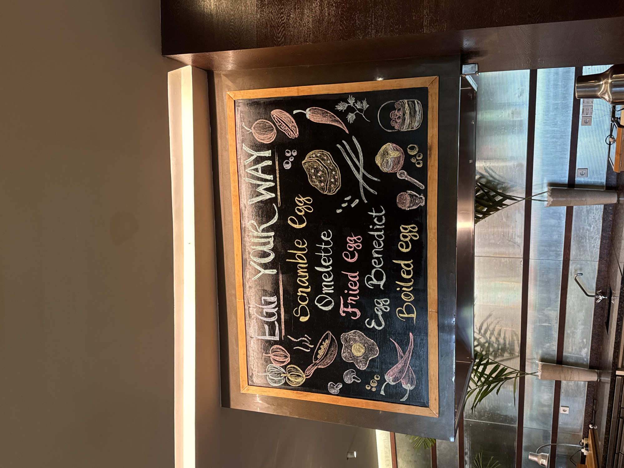 The image depicts a close-up view of a blackboard menu titled "Egg Your Way" at a food establishment in Kuta Selatan, Indonesia. The menu is written in chalk with colorful drawings of various ingredients and egg preparations. Items listed include Scramble, Omelette, Fried Egg, Egg Benedict, and Boiled egg. The blackboard is framed in wood and hangs above a kitchen counter with stainless steel appliances and decorative plants in vases. The overall setting appears to be a hotel or restaurant kitchen during the daytime.