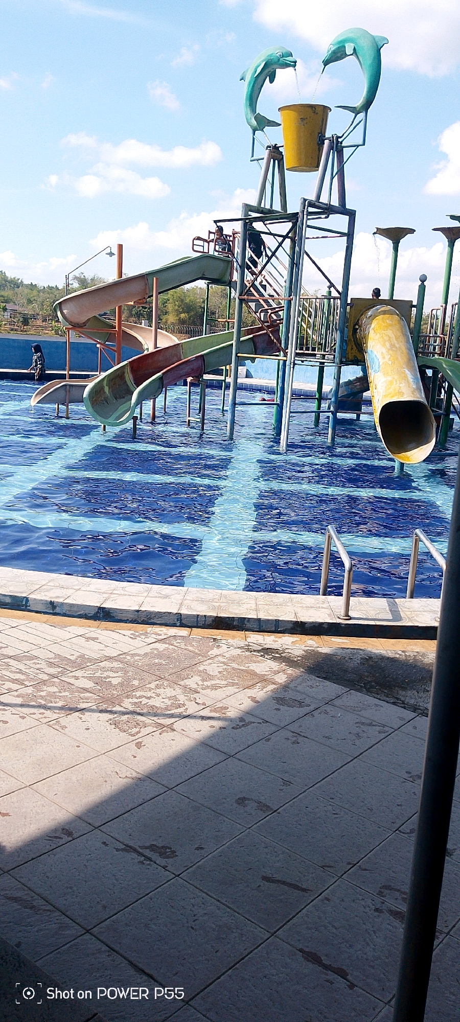 A bright, sunny day at a water park with a blue, tiled swimming pool. In the center of the image, a multi-level water slide structure stands, featuring several slides of varying colors and shapes, including a yellow tube slide and green slides. At the top of the structure, two teal dolphin statues spout water into a yellow bucket. Several people are visible, including one person in a blue swimsuit and headscarf standing at the edge of the pool, and a few individuals in the background near the slides. The pool's surface is rippled, and a tiled walkway surrounds it, with some wet patches and shadows. The background shows greenery and hills under a sky with scattered clouds.