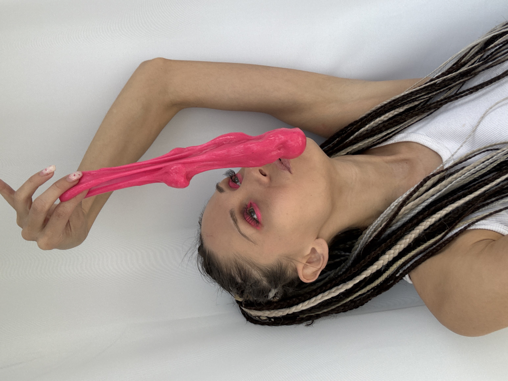 A woman with brown and blonde braided hair, wearing a white tank top, lies against a white background. She holds a piece of pink slime that stretches from her hand to her face, covering her nose. She wears pink eyeliner. There is no indication that the image was taken in Rusanovo, Russia.