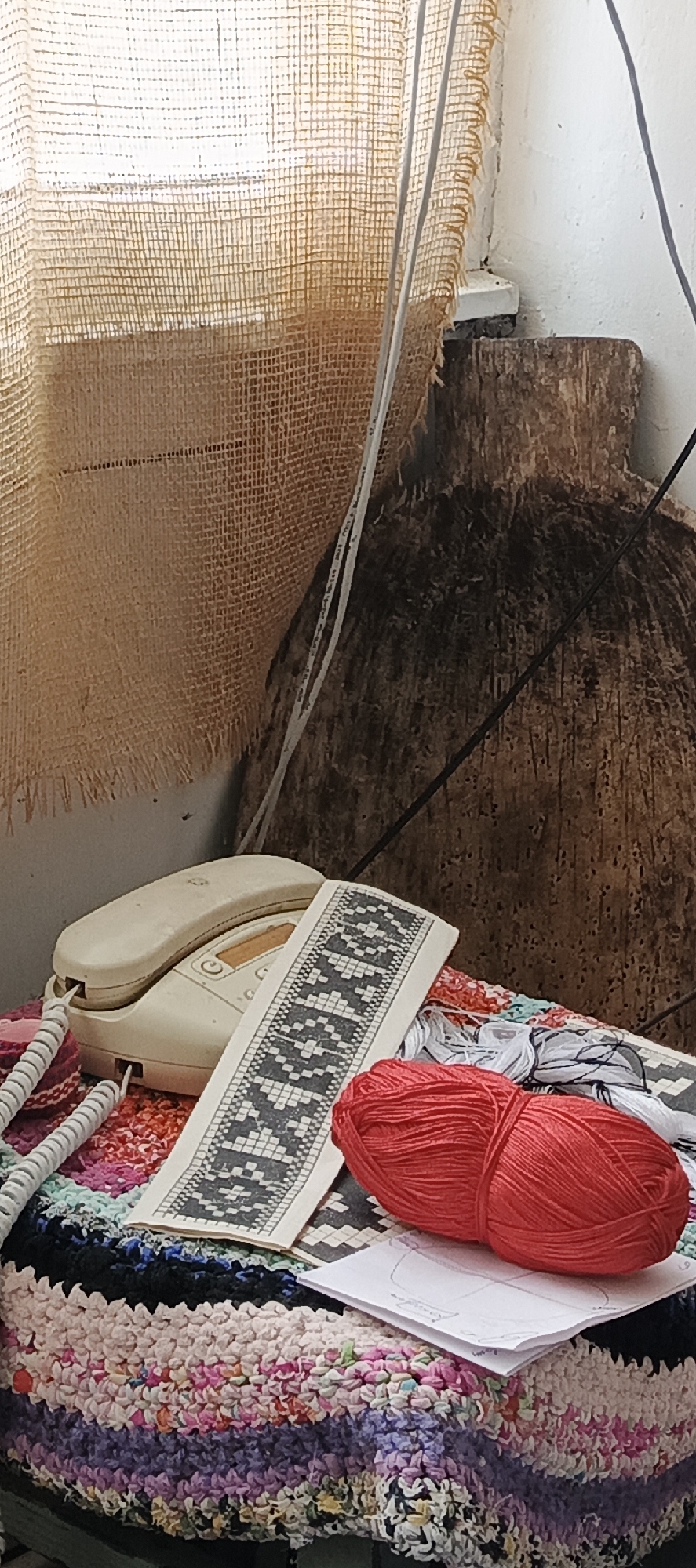 The image shows a still life scene. A landline telephone sits on a colorful, textured fabric. Adjacent to the telephone is a paper pattern with a geometric design. A ball of red yarn rests nearby. The background includes a burlap curtain and a dark, wooden object. The specific location within Кемуль, Russia, cannot be determined from this image alone.