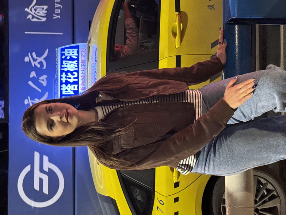 A young adult female is depicted in the foreground, seated or leaning on a blue urban barrier. She faces slightly right of the viewer, exhibiting a subtle smile. She wears a dark brown cropped jacket over a white and dark-striped long-sleeved shirt, paired with light blue jeans. Her fingernails are painted dark blue with lighter accents. Her left hand rests on the blue barrier, while her right hand is placed on her left thigh.
Behind the female subject, a bright yellow taxi cab is parked. The taxi's rooftop features an illuminated white sign displaying Chinese characters "塔花椒油" (Tower Prickly Ash Oil) and "新世纪线上线下超市可购" (New Century Online and Offline Supermarket available). The number "76" is visible on the taxi's rear passenger door. A reflection in the driver's side window reveals a male individual, presumably the driver, with hands on the steering wheel, wearing a light-colored shirt.
In the background, behind the taxi, a large illuminated blue sign displays a white circular logo, likely a stylized 'GT' or 'JT', alongside Chinese characters including "公交" (Public Transport) and "Yuyu." This signage confirms the setting as an urban environment within Yuzhong District, China. The scene is artificially lit, indicating a nighttime or low-light condition.