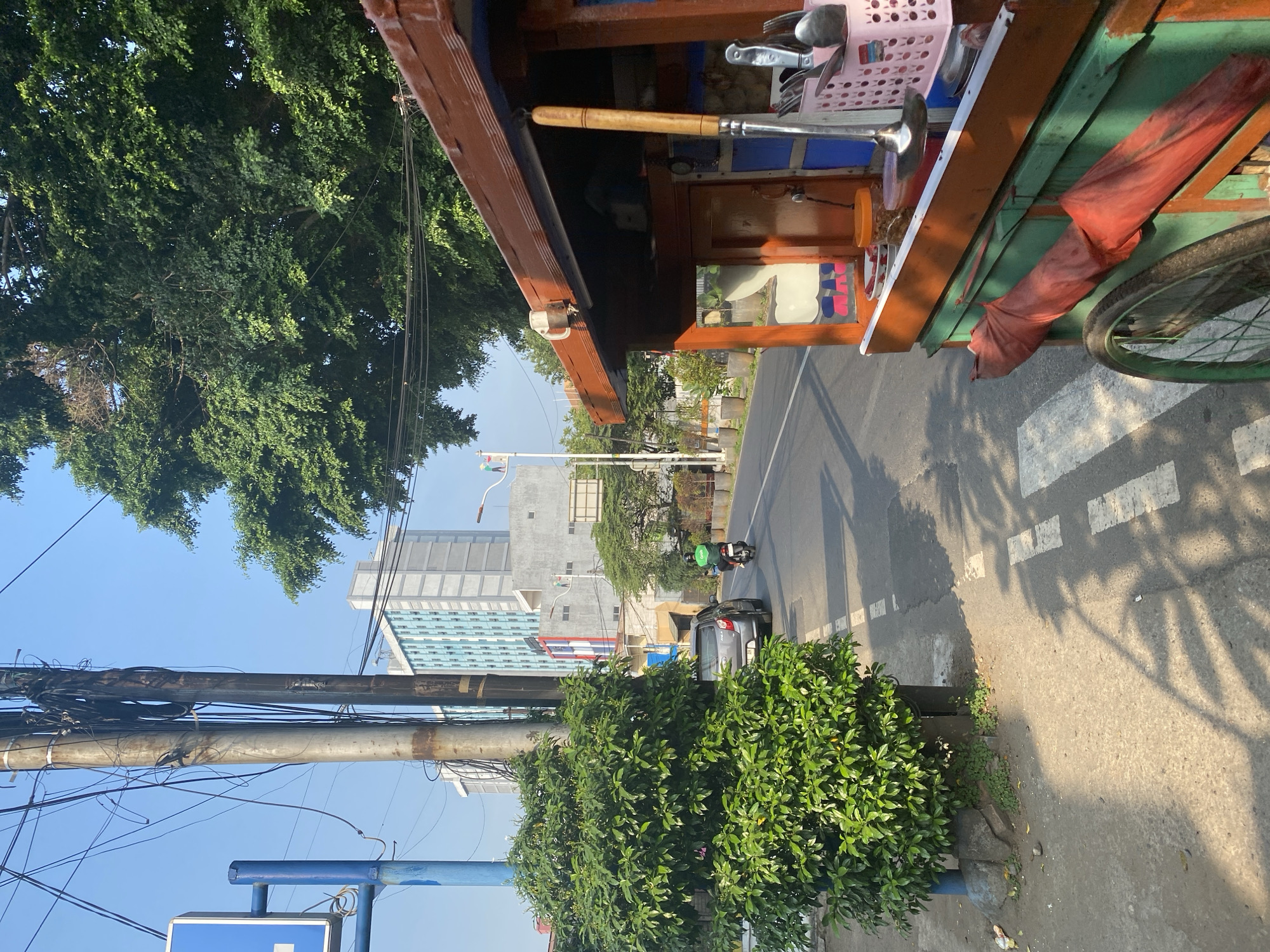 The image depicts a street scene in East Jakarta, Indonesia, on a sunny day. In the foreground, a colorful food cart with a wooden roof and green base is parked on the right side of the frame. Various utensils, including ladles and spoons in a pink basket, are visible on the cart. A red cloth is draped over the front of the cart.
The street runs horizontally across the middle of the image, with a gray car and a motorcycle rider wearing a green jacket visible on it. A white dashed line marks the edge of the lane. On the left side of the street, a lush green tree stands near a utility pole with numerous tangled wires. Behind the tree, two tall buildings, one with a light blue facade and the other with a gray facade, rise into the clear blue sky. The ground is paved and shows shadows cast by the foliage and overhead wires, creating a dappled effect. The scene suggests a busy urban environment with a mix of commercial activity and residential buildings.