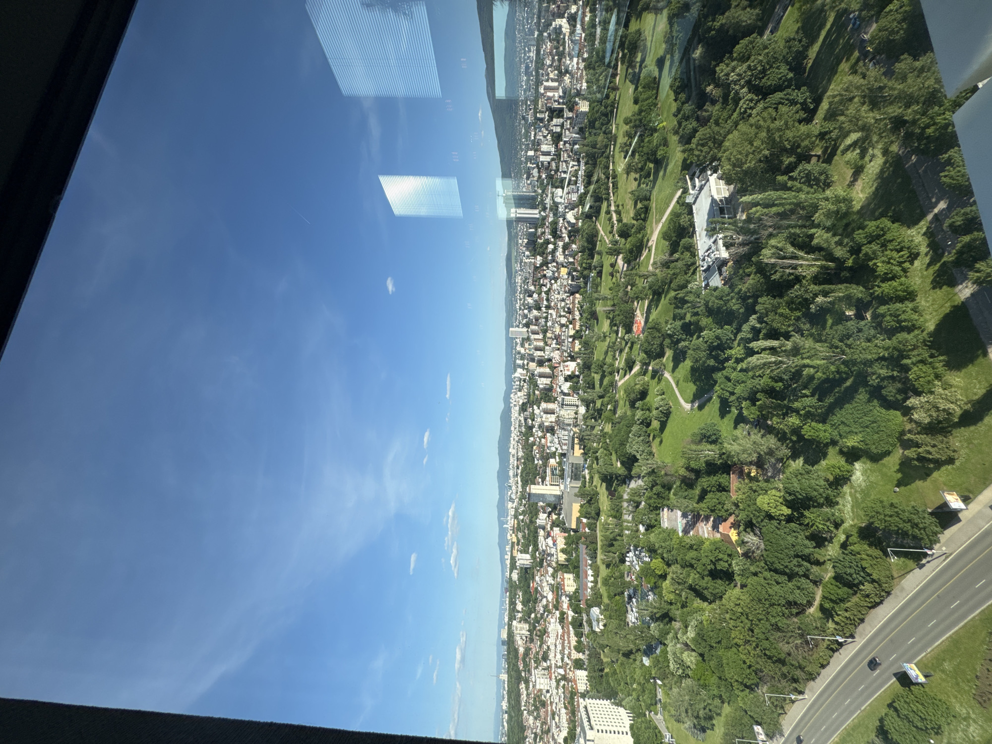 The image shows an aerial view of Sofia, Bulgaria, through a window, reflecting part of the window frame and a building in the sky. The city is visible below, featuring a mix of buildings and green spaces. A major road runs along the edge of a large park.
