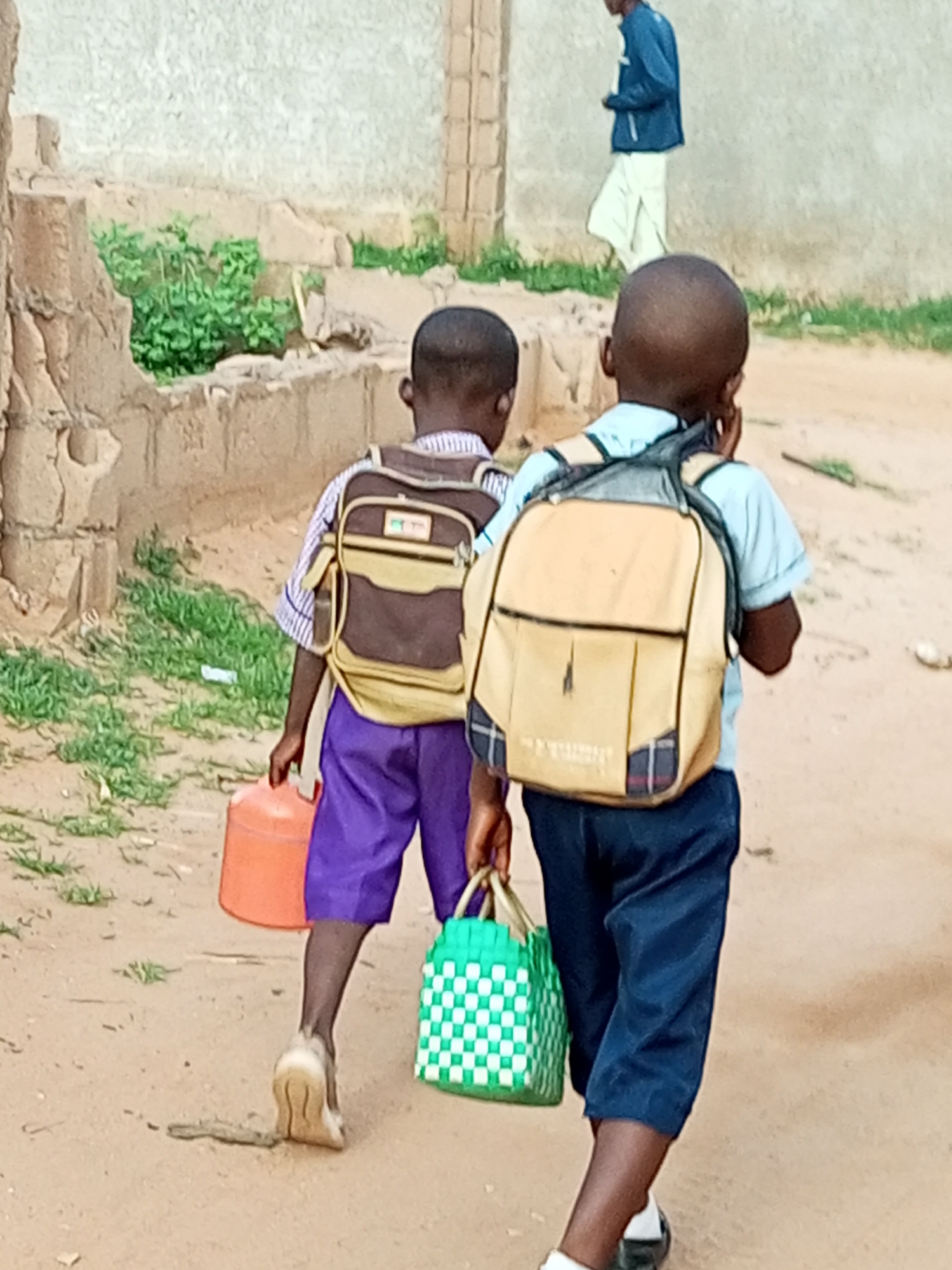 In Tumfure, Nigeria, two young boys walk away from the camera on a dirt path. The boy on the left wears a blue and white striped shirt, purple shorts, and carries a brown and tan backpack and an orange plastic container. The boy on the right wears a light blue short-sleeved shirt, dark blue pants, and carries a light tan backpack and a green and white woven bag. In the background, another person walks away wearing a blue jacket and white pants. The environment consists of rough, textured walls and sparse vegetation.