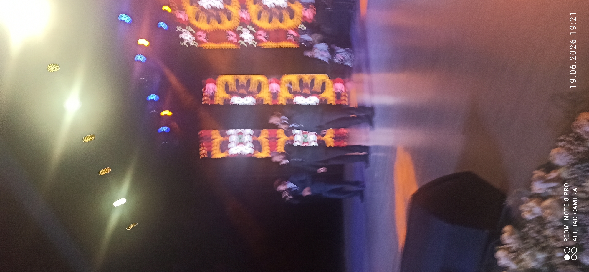 A group of four male performers, dressed in dark suits, are singing on a stage with a large, colorful LED screen behind them. The screen displays a vibrant, abstract pattern of yellow, orange, red, and white. Three of the performers are in the foreground, singing into microphones. One performer, slightly to the left, is looking down while singing. Another performer is in the background, also singing. The stage floor is a light gray, reflecting the stage lights. In the upper part of the image, several rows of circular lights are visible, some glowing yellow and blue. There is also a bright light source in the upper right corner, illuminating the scene. A portion of a black speaker is visible in the lower left corner. The bottom right corner displays a date and time stamp: "19.06.2026 19:21", and a watermark that reads "REDMI NOTE 8 PRO AI QUAD CAMERA". The image appears to be taken from a low angle.