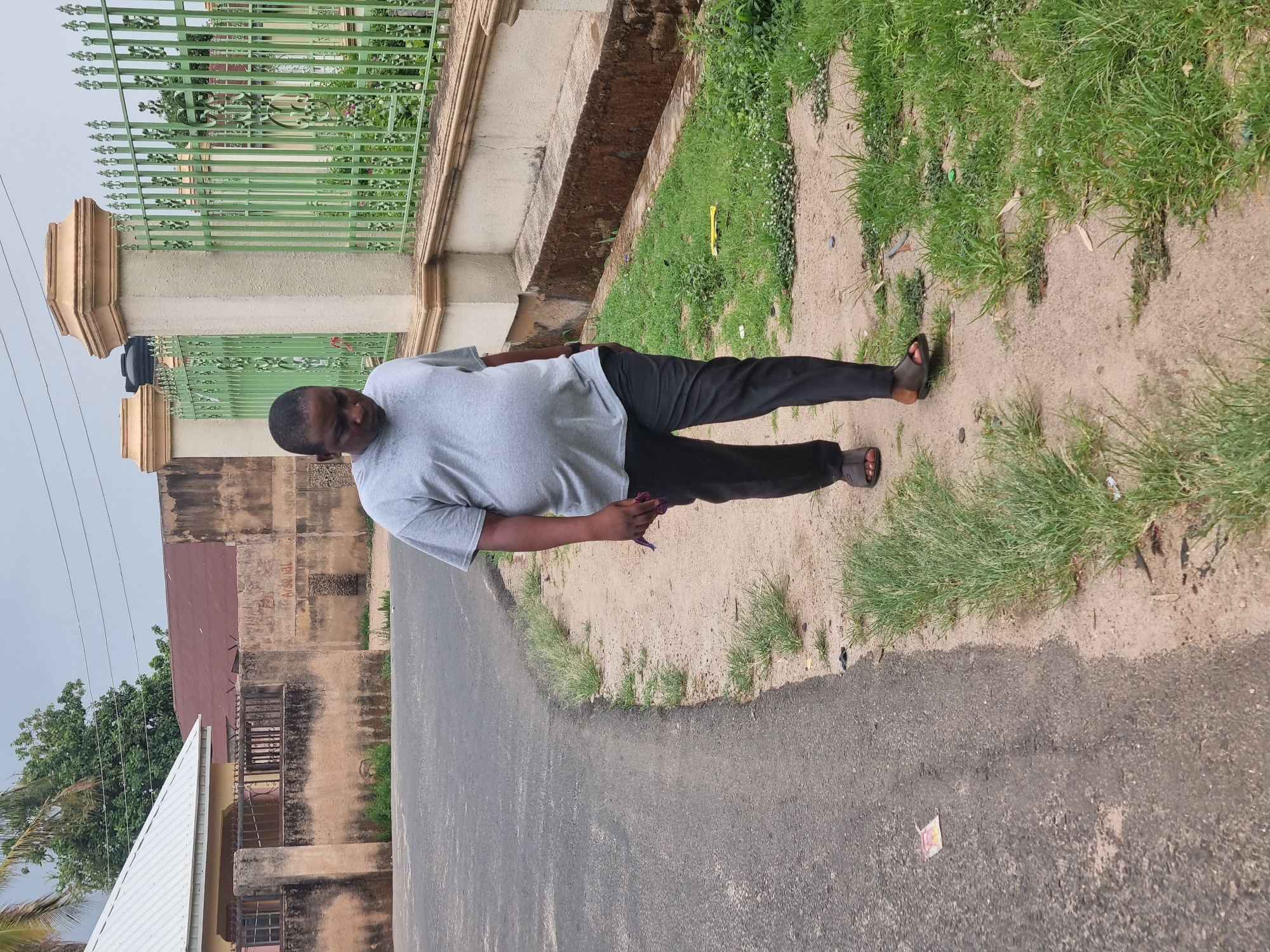 A person stands on a street adjacent to a dirt shoulder and a patch of grass. The person wears a gray t-shirt, dark pants, and sandals. A building with a green metal fence borders the street. The environment suggests an urban area, potentially in Jos, Nigeria. The scene is captured in daylight.