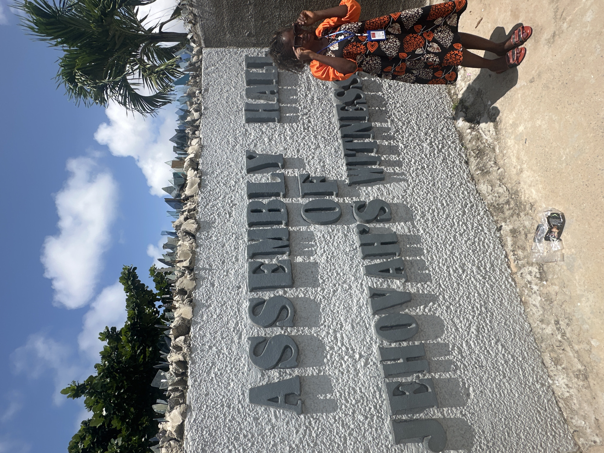 The image captures an outdoor scene at the Assembly Hall of Jehovah's Witnesses in Itoga, Nigeria. A young girl, wearing sunglasses, an orange shirt, and a patterned dress, stands to the right of a textured white wall. The wall features prominent grey lettering spelling out "ASSEMBLY HALL OF JEHOVAH'S WITNESSES." The top of the wall is lined with what appear to be shards of glass or broken material, set into a concrete barrier, likely for security. Behind the wall, lush green trees and a palm tree are visible against a bright blue sky with scattered white clouds. The ground in front of the wall is a rough, light-colored concrete or dirt surface, with some debris, including a crumpled plastic bag, on the lower right. The sunlight casts distinct shadows from the lettering and the girl onto the wall and ground, indicating it is a sunny day.