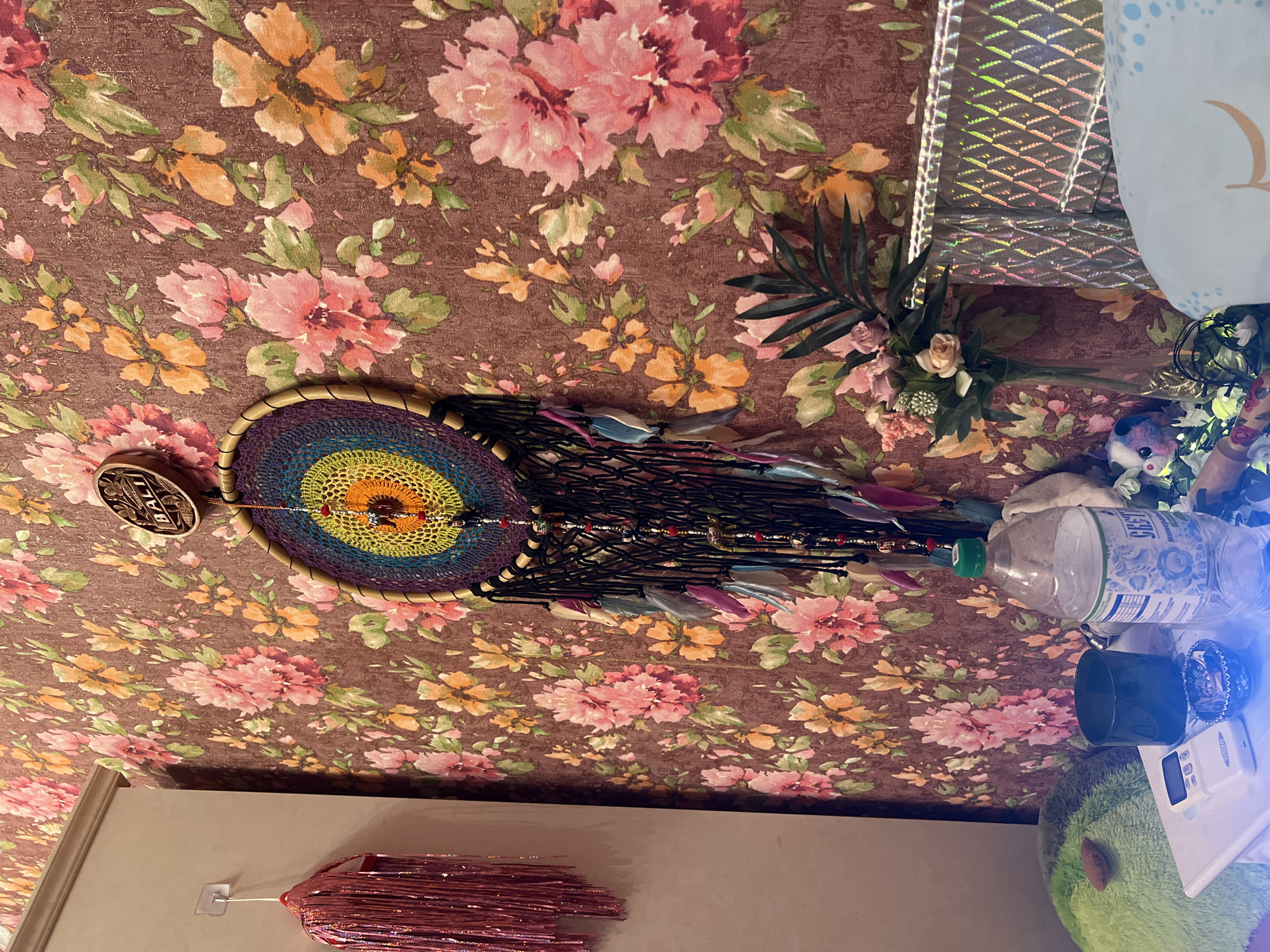 A colorful dreamcatcher hangs on a floral wallpapered wall. To the left, a pink tinsel fringe hangs from a hook. On a white surface below, a plastic water bottle, a small black cup, a white electronic device, and a decorative glass bowl are present. In the background, a vase with artificial flowers and a shiny textured object are visible.