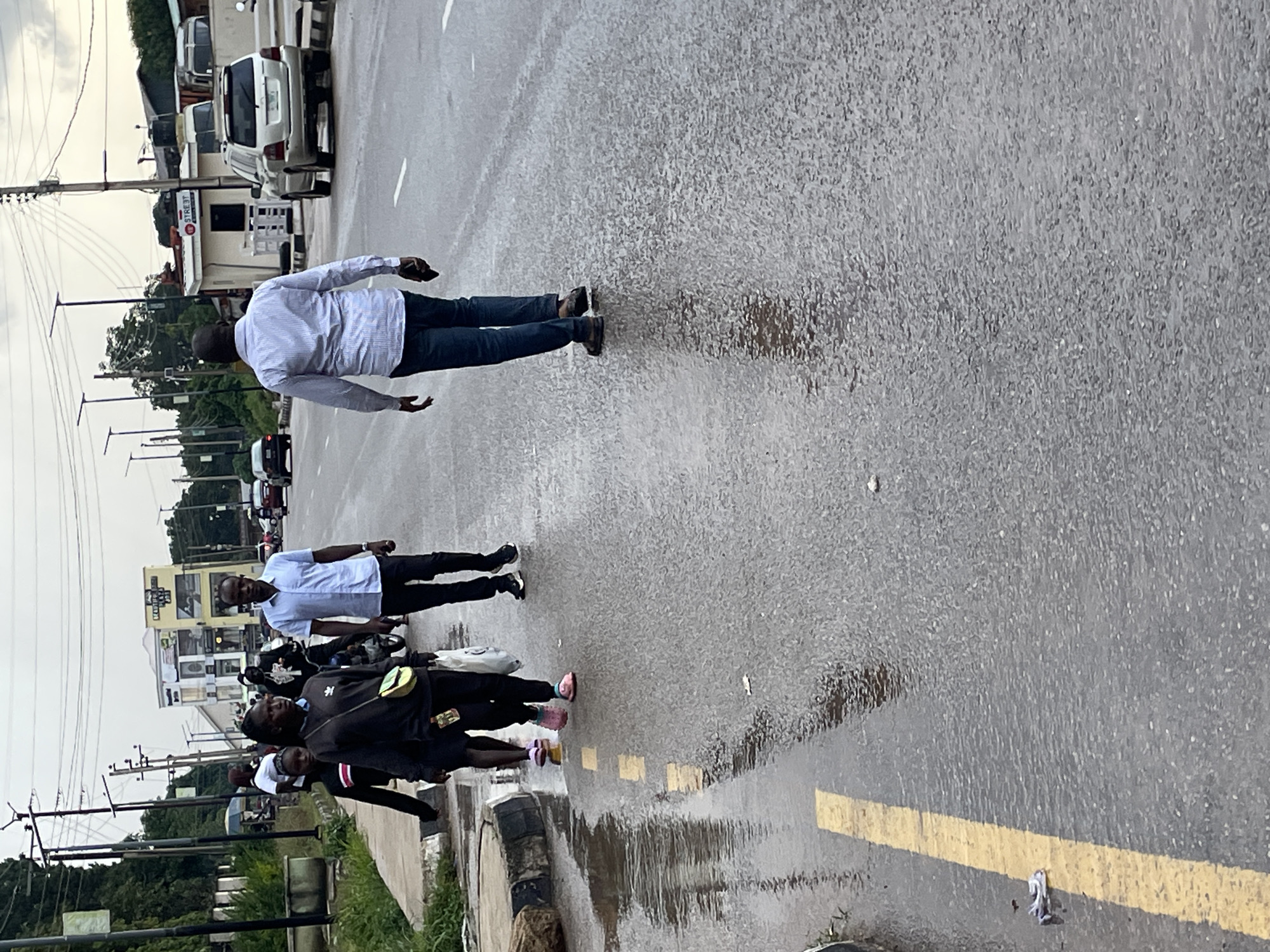 On a cloudy day, a group of people walk on a wet asphalt road in Jos, Nigeria. A man in a light blue shirt and dark pants walks away from the camera towards the right side of the frame, holding a phone in his right hand. To his left, a man in a striped shirt and dark pants walks in the same direction. To the left of that man, a group of three women walk on the side of the road, closest to the camera. The woman in the middle wears a black long-sleeved shirt and a yellow crossbody bag. Her feet are wet, and she is holding a plastic bag in her right hand. A woman in a black dress and pink shoes walks behind her. A third woman, wearing a white shirt, is partially visible behind the woman in the black dress. The road has puddles of water, indicating recent rain. Buildings and utility poles are visible in the background.