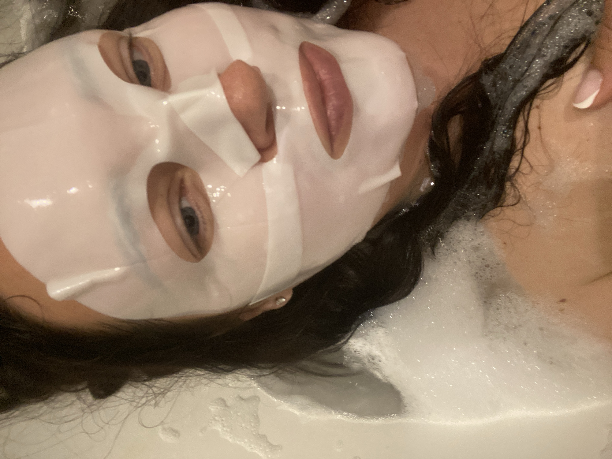 A person with a white sheet mask on their face is partially submerged in a bathtub filled with white bubbles. Their dark hair is wet and mixed with the bubbles. The person's eyes and mouth are visible through the mask. The location is not discernible from the image.