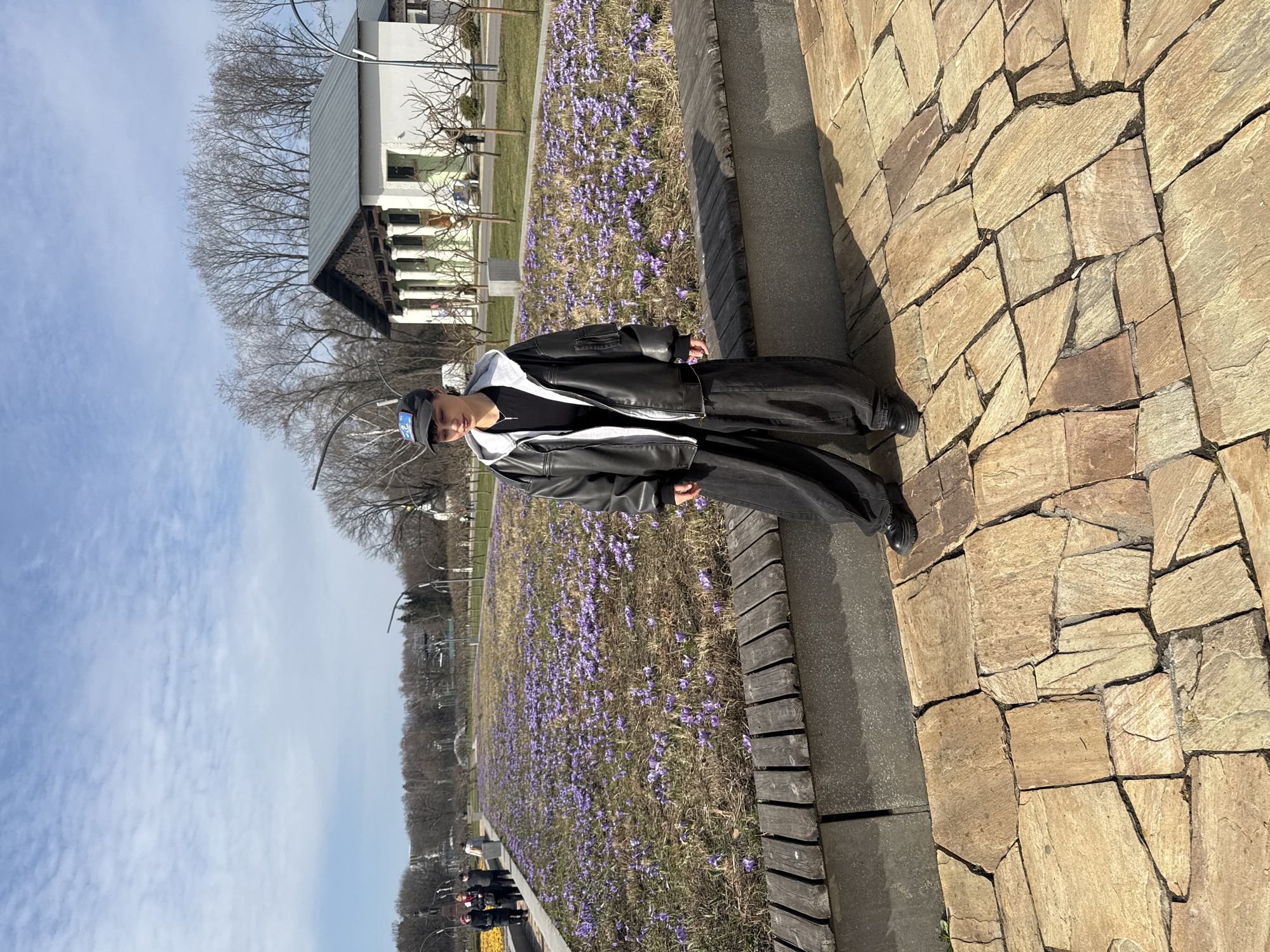 A person stands on a brick walkway next to a flower bed filled with purple flowers. The person is wearing a black jacket with white trim, black pants, and a cap. A building with white columns can be seen in the background, along with trees and a blue sky with some clouds. Based on the style of the building and landscape, the image is likely taken in a park setting in Moscow, Russia.