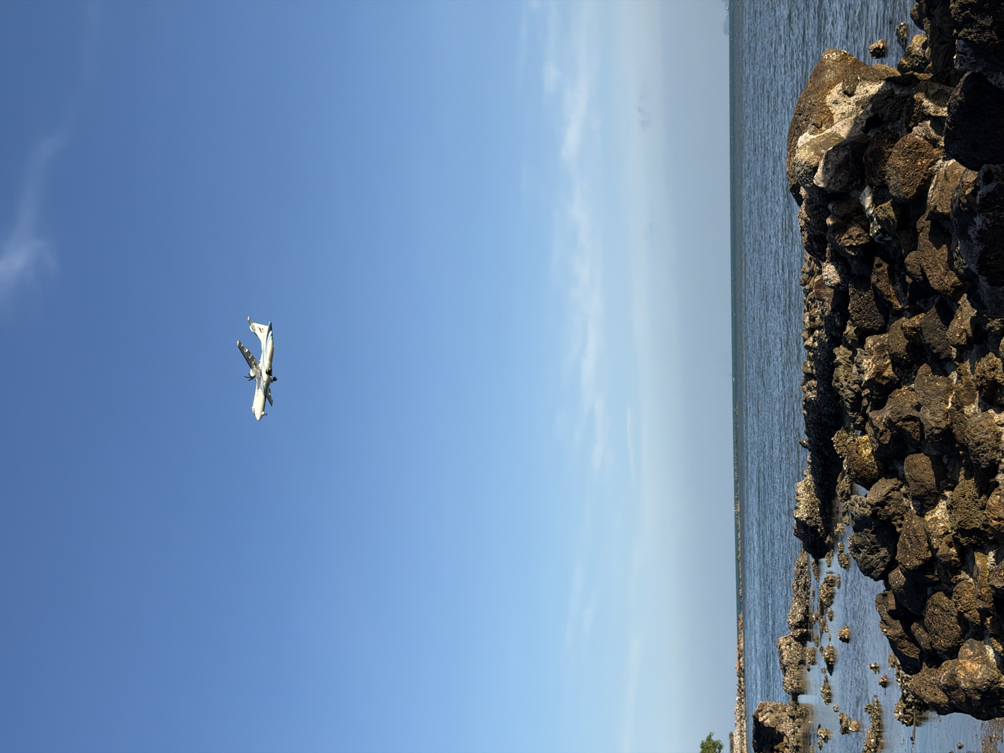 A white twin-engine turboprop aircraft with visible propellers flies across a clear blue sky above a rocky coastline. The aircraft is angled slightly downwards, suggesting it is in the process of landing. The foreground is dominated by dark, rough-textured rocks of various sizes, some submerged in the calm, blue water of the sea. In the background, the sea extends to the horizon, with a faint line of clouds visible near the surface. A distant, dark shape on the horizon might be a ship. A single green tree is visible on the far left of the frame. The scene depicts a coastal area in Ko Samui District, Thailand, under a bright, sunny sky.