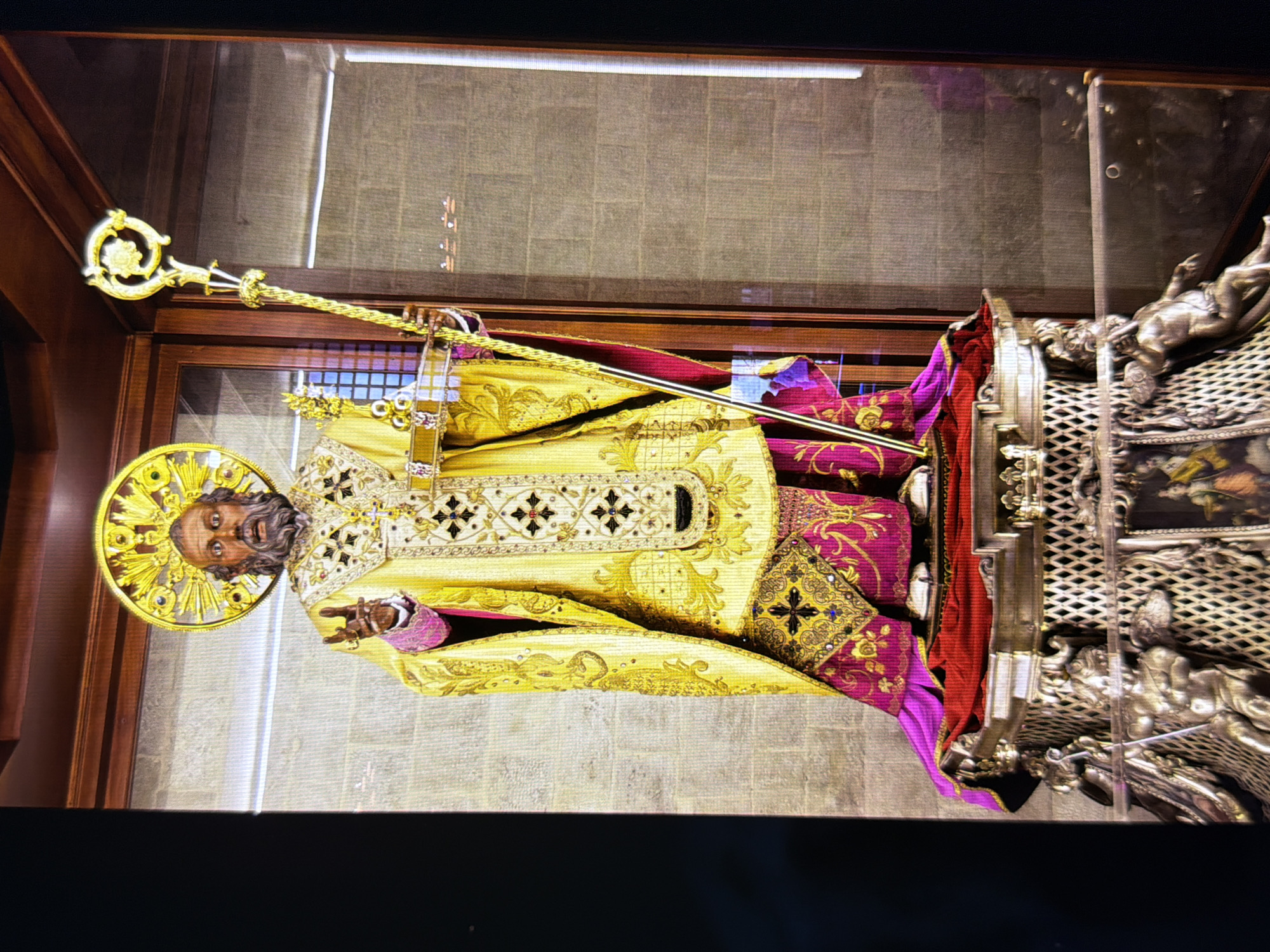 The image shows a statue of a saint, likely St. Nicholas, dressed in ornate religious vestments. The statue is gold and purple, with intricate embroidery. It stands on a pedestal adorned with metal figures. The background appears to be a museum or church interior, with brick walls and wooden display cases. The statue is holding a staff in its right hand and a book or reliquary in its left. The halo around the saint's head is also gold and decorated. The overall impression is of a valuable and historic religious artifact.
This image is not located in Kyiv, Ukraine.