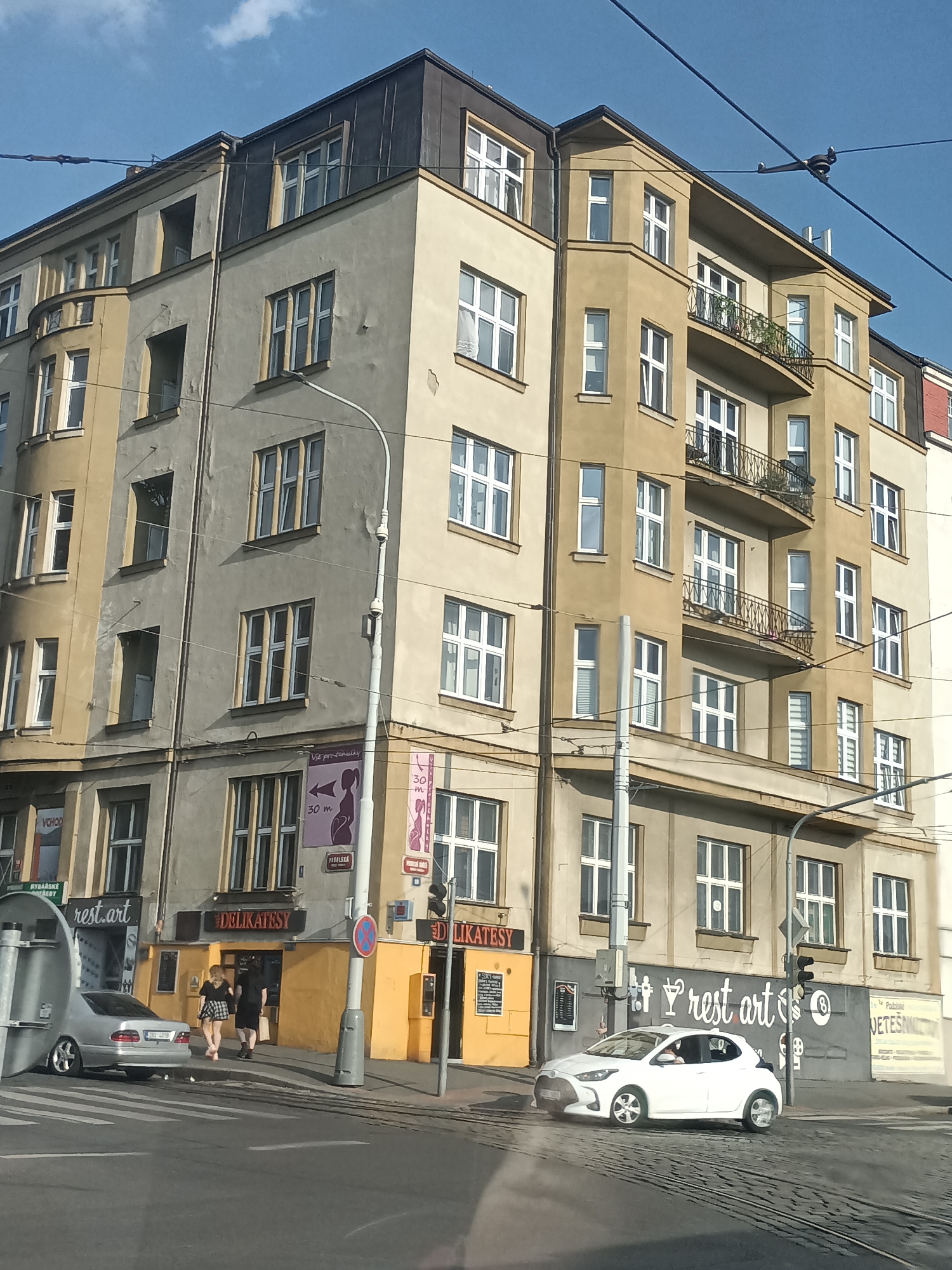 This is a medium-angle, eye-level outdoor shot of a mixed-use building located in Prague, Czechia, on a clear day with a blue sky. The building is tall and appears to be an older apartment or office building with a yellowish-tan facade and several windows on each floor. The corner of the building is rounded and features balconies. A street runs in front of the building, with a crosswalk and tram tracks visible.
In the foreground, a silver Mercedes-Benz sedan is parked on the right side of the street, and a white Toyota Yaris is driving across the intersection on the left. Two pedestrians, a woman with blonde hair and a man with dark hair, are walking on the sidewalk in front of a store with a yellow facade. The store has a sign that reads "DELIKATESY".
Other visible details include overhead power lines, a street lamp, a traffic light, and several posters and signs on the building, including one with a silhouette of a pregnant woman. The architectural style suggests the building is from the early 20th century.
