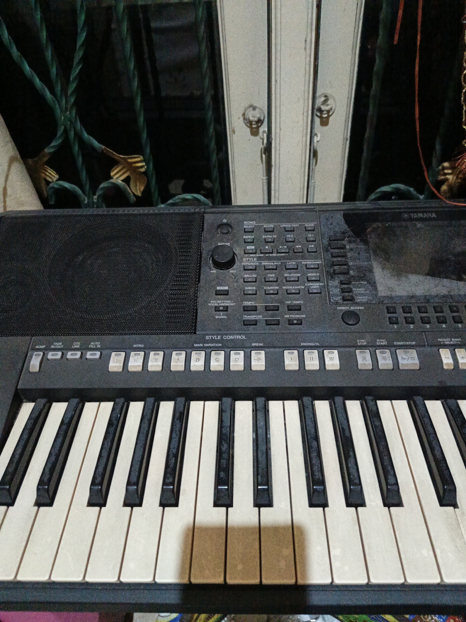 An electronic keyboard, specifically a Yamaha brand, is the primary subject in this indoor image. The keyboard is positioned horizontally, filling the majority of the frame, with its keys and control panel clearly visible. The control panel features numerous buttons, knobs, and a display screen. The keys are a mix of white and black, standard for a keyboard instrument. Behind the keyboard, a dark, textured surface is visible, possibly a wall or fabric, with a green, twisted metal grate or decorative element overlaying it. There are also hints of vertical white surfaces with metallic accents that resemble doors or shutters in the background, suggesting an indoor environment with some architectural features. The lighting appears to be artificial and somewhat dim, casting faint shadows. The location is identified as Karangpakel, Indonesia. No people are present in the image.