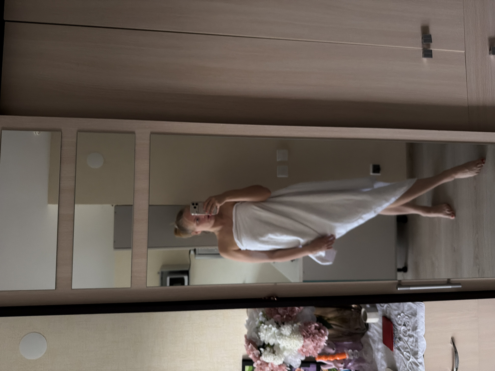 A mirror reflects a woman wrapped in a white towel, holding a smartphone and taking a selfie. The background includes a room interior with a microwave, a cabinet, a bed, and decorative flowers. The reflection suggests the photograph was taken in a bedroom. There is no information to identify this location as being in Izhevsk, Russia.