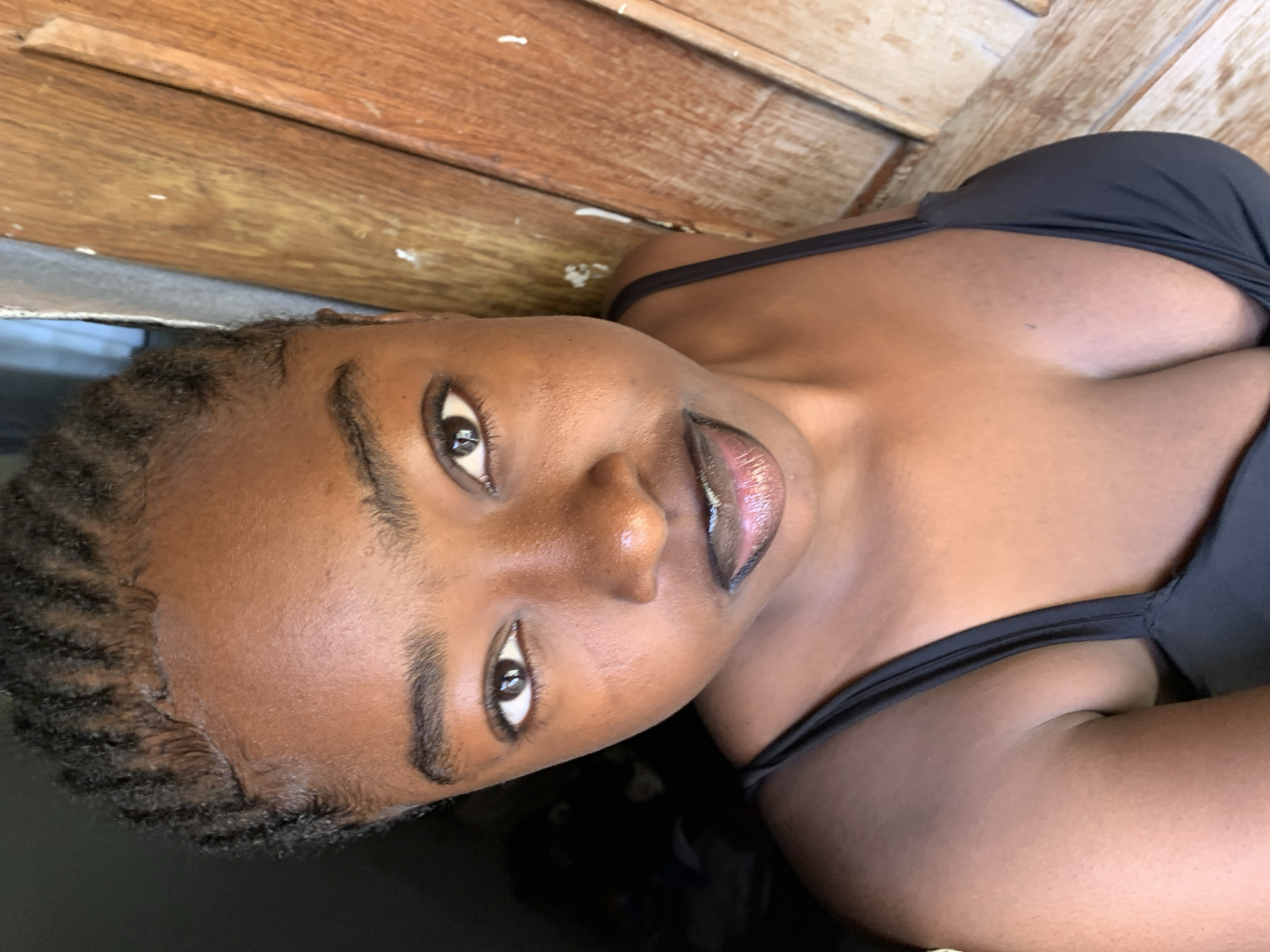 A young Black woman with braided hair and a black top is shown in a close-up, low-angle shot. She is looking directly at the camera, with her eyes open. Her lips are slightly parted and appear to be wearing a dark lipstick. The background is divided into two sections: on the left, a dark, shadowy area, and on the right, a wooden door or wall with vertical planks. The lighting suggests she is outdoors or near an open doorway, possibly in Rafin Par, Nigeria, as indicated by the wooden structure common in such environments. The overall impression is a casual, personal photograph.