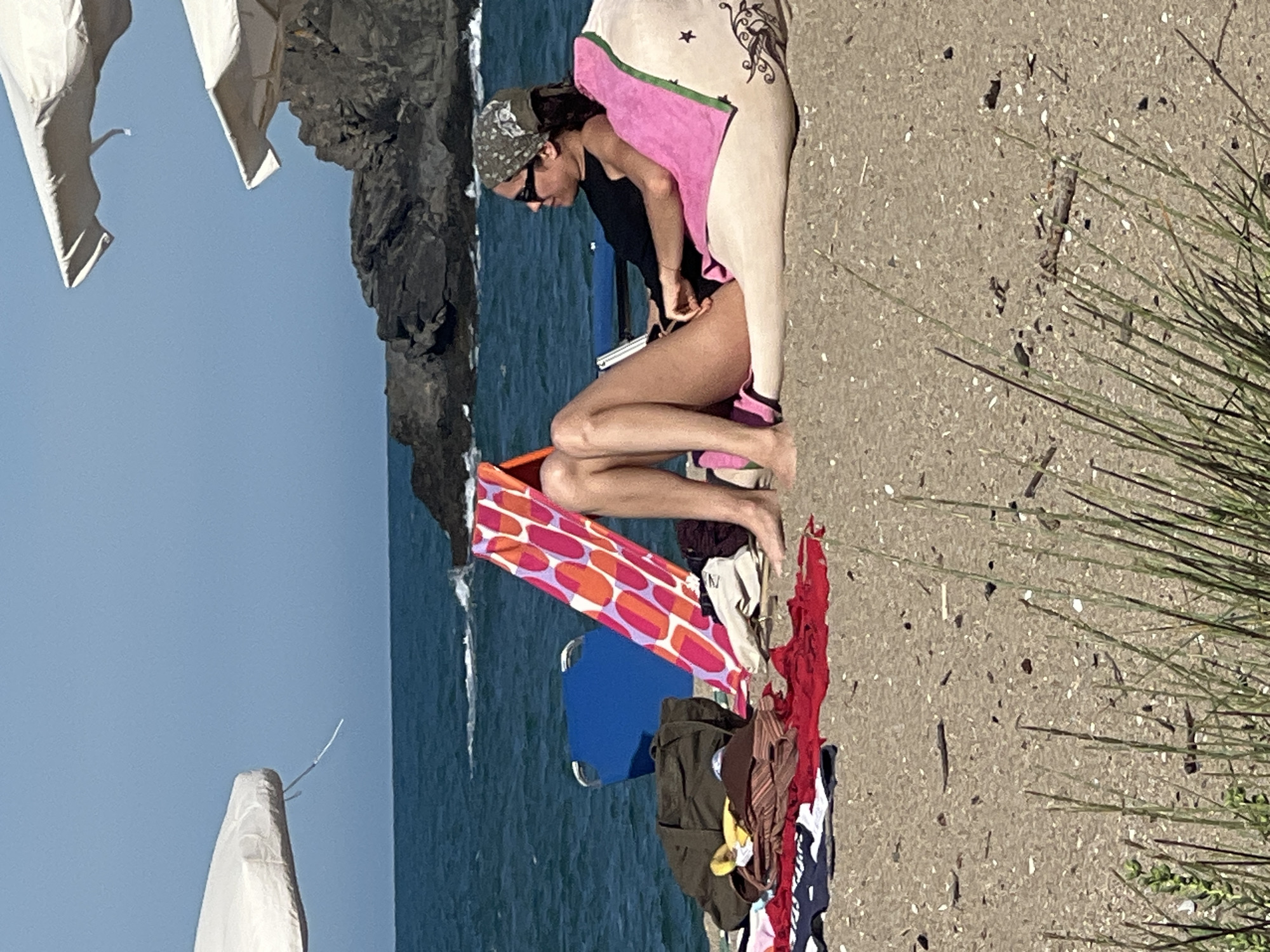 A woman is relaxing on a beanbag on a sandy beach, facing to the right. She is wearing a black tank top and a bandana. The beanbag is pink and white with some decorative elements. In front of her, there are various beach items, including towels, a bag, and a foldable blue chair. A brightly colored towel with an orange and pink pattern stands upright behind some of the items. The sea is visible in the background with some rocks. The sky is clear and blue. The location is the beach in Sinemorets, Bulgaria.