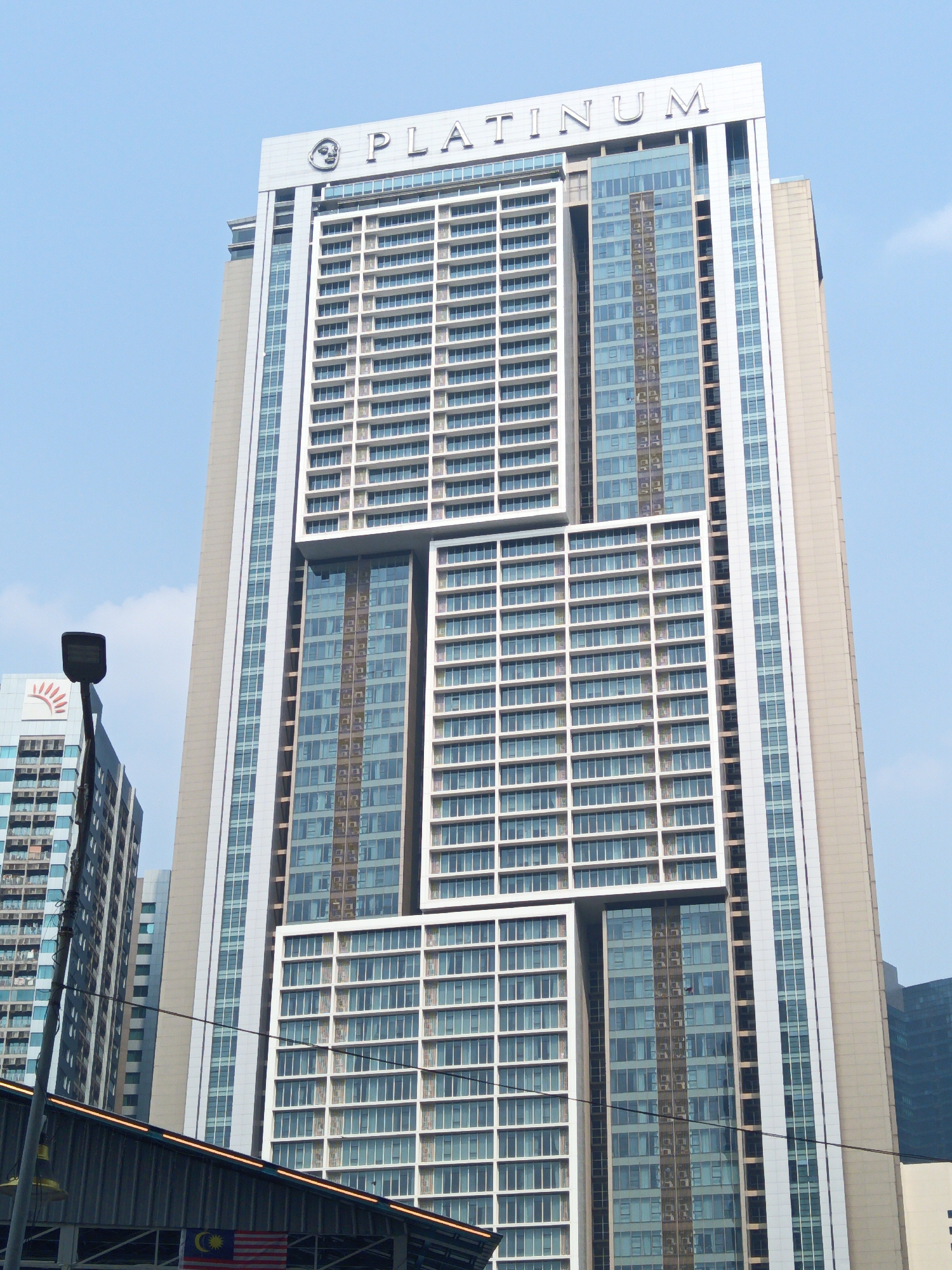 The image shows a view of the Platinum Suites building in Kuala Lumpur, Malaysia, under a clear sky. The tall skyscraper is characterized by its modern architecture, featuring a large number of glass-enclosed windows arranged in a grid pattern. The building's facade is predominantly white and glass, with distinct sections of differing window arrangements and proportions, creating a dynamic visual effect. The name "PLATINUM" is prominently displayed in large, silver letters on the top of the building.
In the foreground, a portion of a building or structure is visible, with a Malaysian flag hanging from it. A lamppost with a modern, rectangular light fixture is also present, partially obscuring the view of another building to the left, which has a logo of a rising sun. The overall scene depicts an urban environment with a focus on contemporary architecture and city infrastructure.