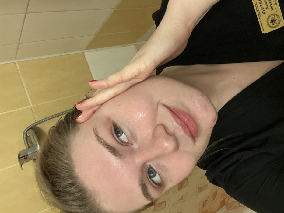 A woman with wet hair poses, touching her forehead with a hand displaying red nail polish. She has fair skin, light eyebrows, and light eyes, with glossy lips. She is wearing a black top. The background shows a yellow-tiled wall and a shower head. A badge is visible on her top, featuring text and a small emblem.
