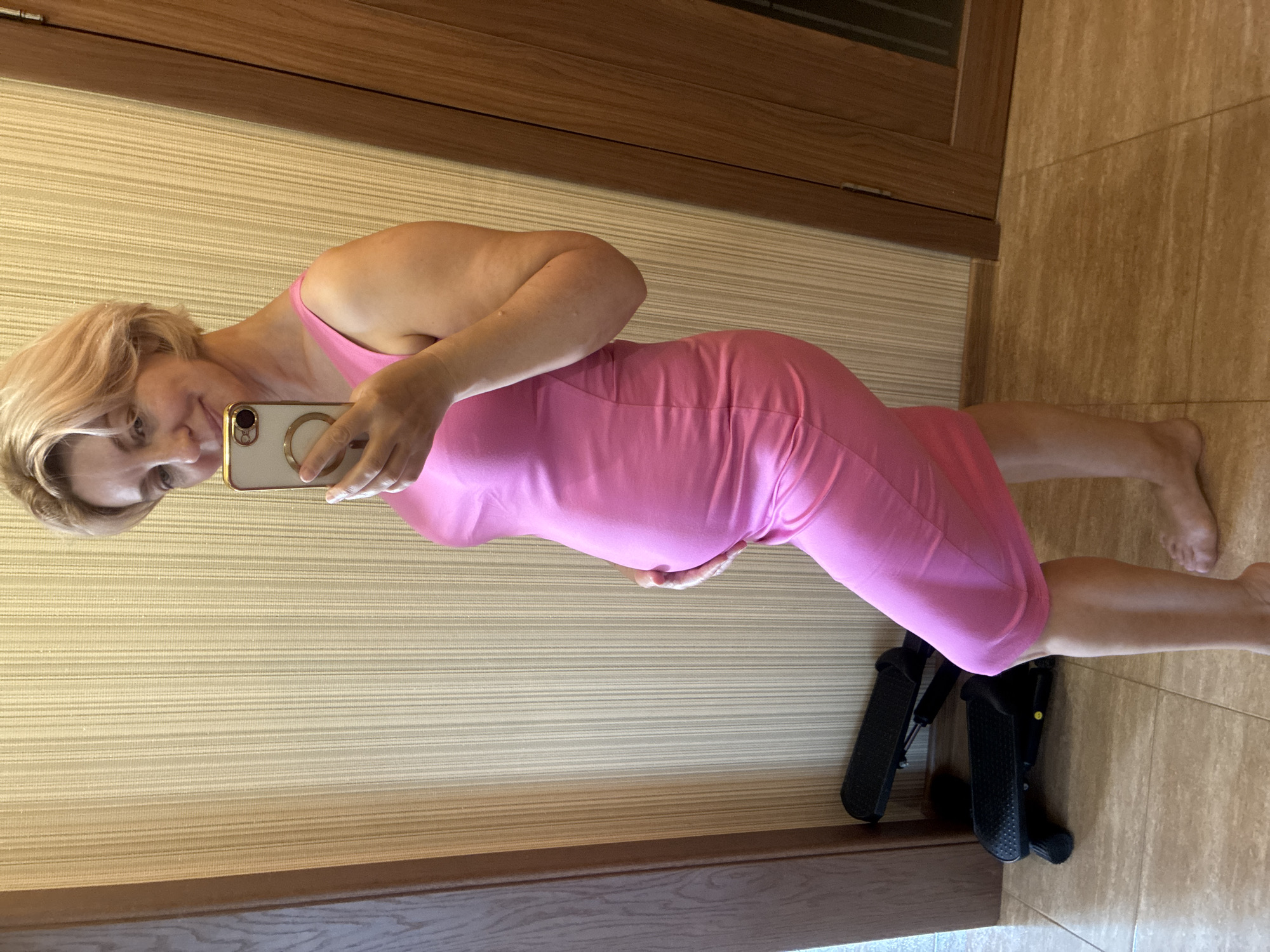 A Caucasian woman, appearing to be in her late 30s or early 40s, stands in profile facing a mirror, taking a selfie. She has short, blonde hair and is wearing a form-fitting, bright pink dress. Her right hand is raised, holding a smartphone, and her left hand rests gently on her abdomen, which appears to be slightly protruding, suggesting pregnancy.
The background is a wall with vertical, textured wallpaper in a light tan color. To the left, a dark wooden door frame is visible, and to the right, another dark wooden door with a glass panel is present. On the floor, which is tiled with light brown and beige square tiles, there are two black exercise steppers positioned in front of the wall. The lighting is a warm, ambient indoor light. There are no indications of the city of Sochi, Russia, within the image itself.