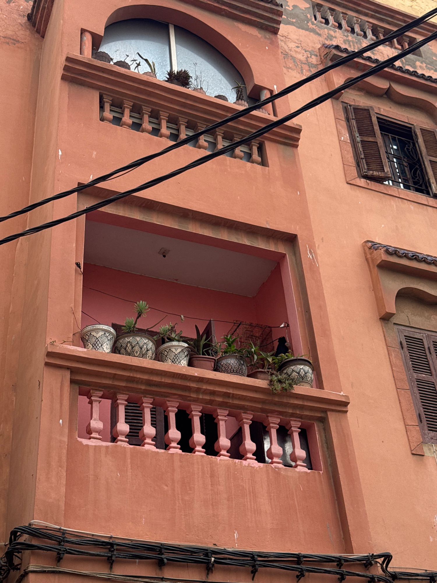 This is a close-up, low-angle, exterior shot of a terracotta-colored building in Essaouira, Morocco. The building features architectural elements characteristic of Moroccan design, including an arched window with plants on the windowsill and a balcony with decorative balusters. Several potted plants are visible on the balcony, some with intricate patterns on the pots. A dark metal railing with a decorative pattern is visible behind the balusters. Black electrical wires are strung across the facade of the building, partially obscuring the view. To the right, a wooden shuttered window is partially open, revealing a dark interior. The building's facade shows signs of age and weathering, with some peeling paint and subtle stains. The overall impression is one of a well-lived-in, traditional urban dwelling.