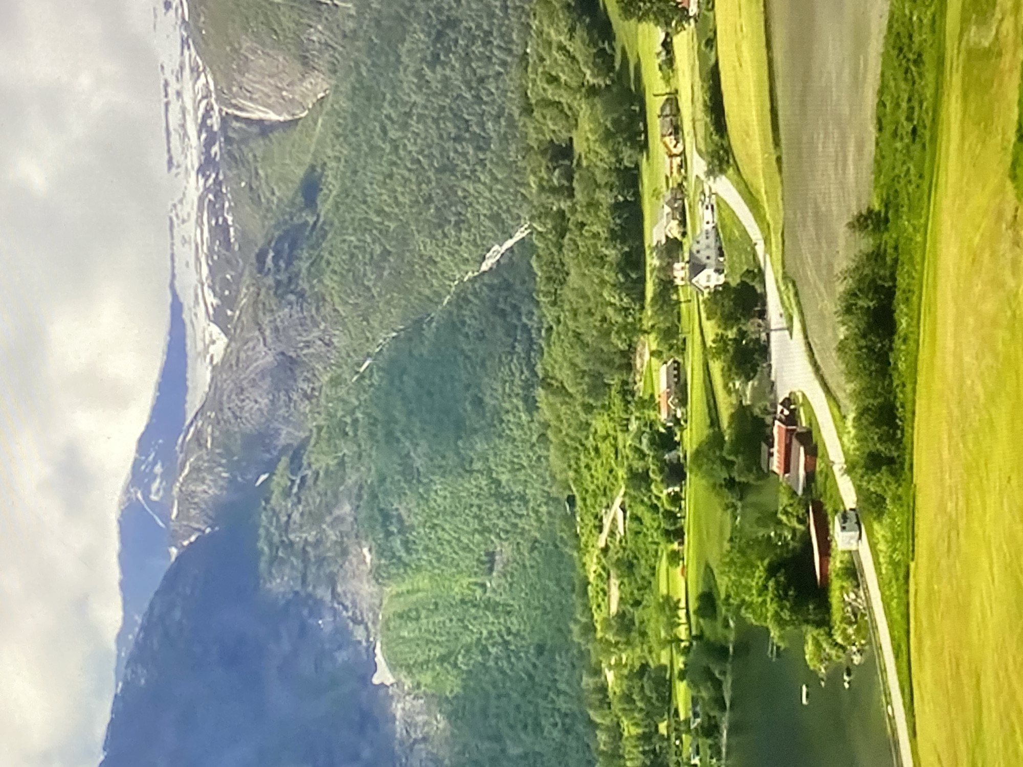 This is an aerial landscape photograph depicting a rural village nestled at the base of a mountain range. The mountains dominate the upper portion of the image, with snow-capped peaks and rugged slopes covered in lush green vegetation. A waterfall is visible cascading down the side of one of the mountains. In the midground, a village of scattered houses with red roofs is surrounded by vibrant green trees and fields. A road winds through the village, and a white camper van is parked on it near a body of water, likely a lake or fjord. The foreground is filled with bright green and yellow fields, suggesting agricultural land. The sky is partly cloudy.
There is no indication that this location is in Ado, Nigeria. The landscape with snow-capped mountains and a fjord-like body of water is not characteristic of Nigeria.