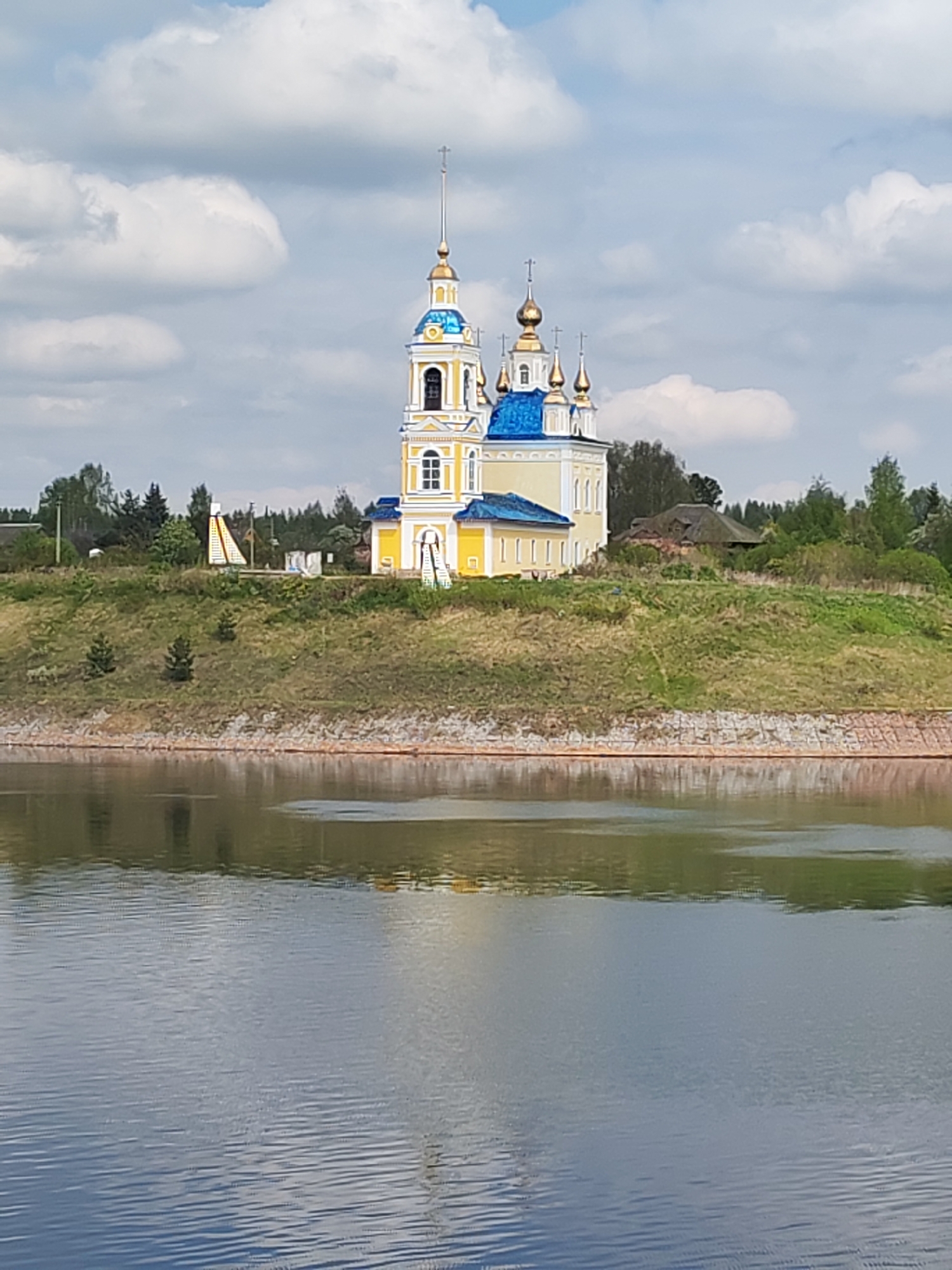 The image shows a church with a blue roof and yellow walls, located on a hill overlooking a body of water. The church has multiple domes with golden spires and a tall bell tower. The sky is partly cloudy. The church is located in Глебово, Russia.