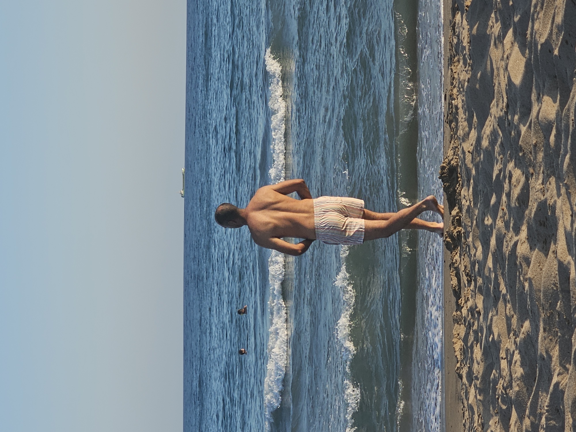 A man in striped swim trunks stands on a sandy beach facing the ocean. The ocean is blue with moderate waves and two people are visible swimming in the distance. The sky is clear and blue. A cargo ship is visible on the horizon. The scene appears to be in the city of Castelldefels, Spain, known for its long sandy beaches and views of the Mediterranean Sea.