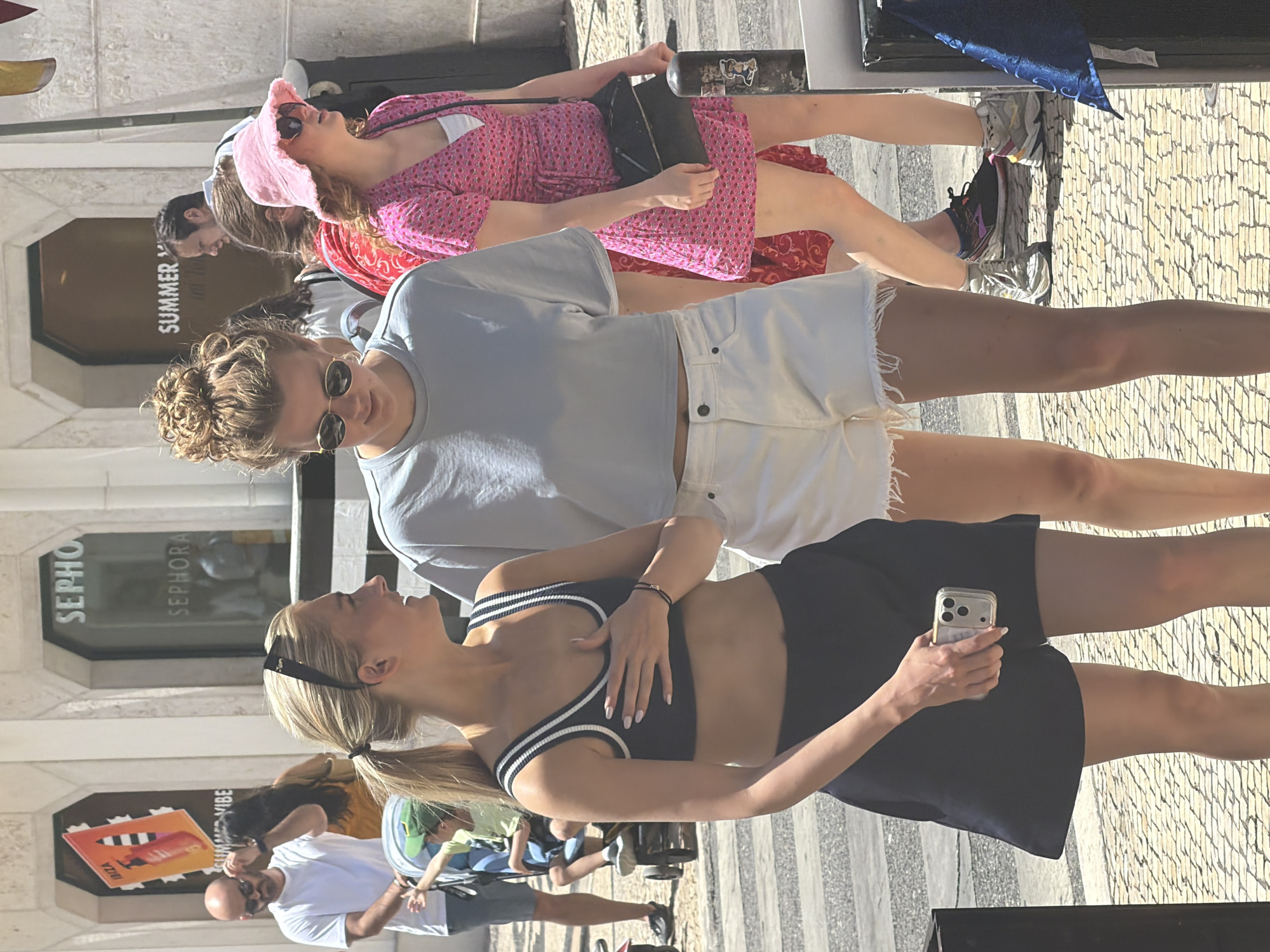 The image captures a street scene in Lisbon, Portugal, on a sunny day. In the foreground, two young women are walking together. The woman on the left has blonde hair pulled back in a ponytail and is wearing a dark athletic crop top with white stripes and dark shorts. She is holding a smartphone in her right hand. The woman on the right has her hair styled in a messy bun and is wearing a light blue t-shirt and white shorts. Both women are wearing sunglasses. Behind them, other pedestrians are visible, including a man in a white t-shirt and shorts pushing a stroller with a child. In the background, shopfronts with signage, including "SEPHORA" and "SUMMER VIBES", are visible, suggesting a commercial area. The ground is paved with traditional Portuguese cobblestones. The overall atmosphere appears casual and lively, typical of a summer day in a European city.
