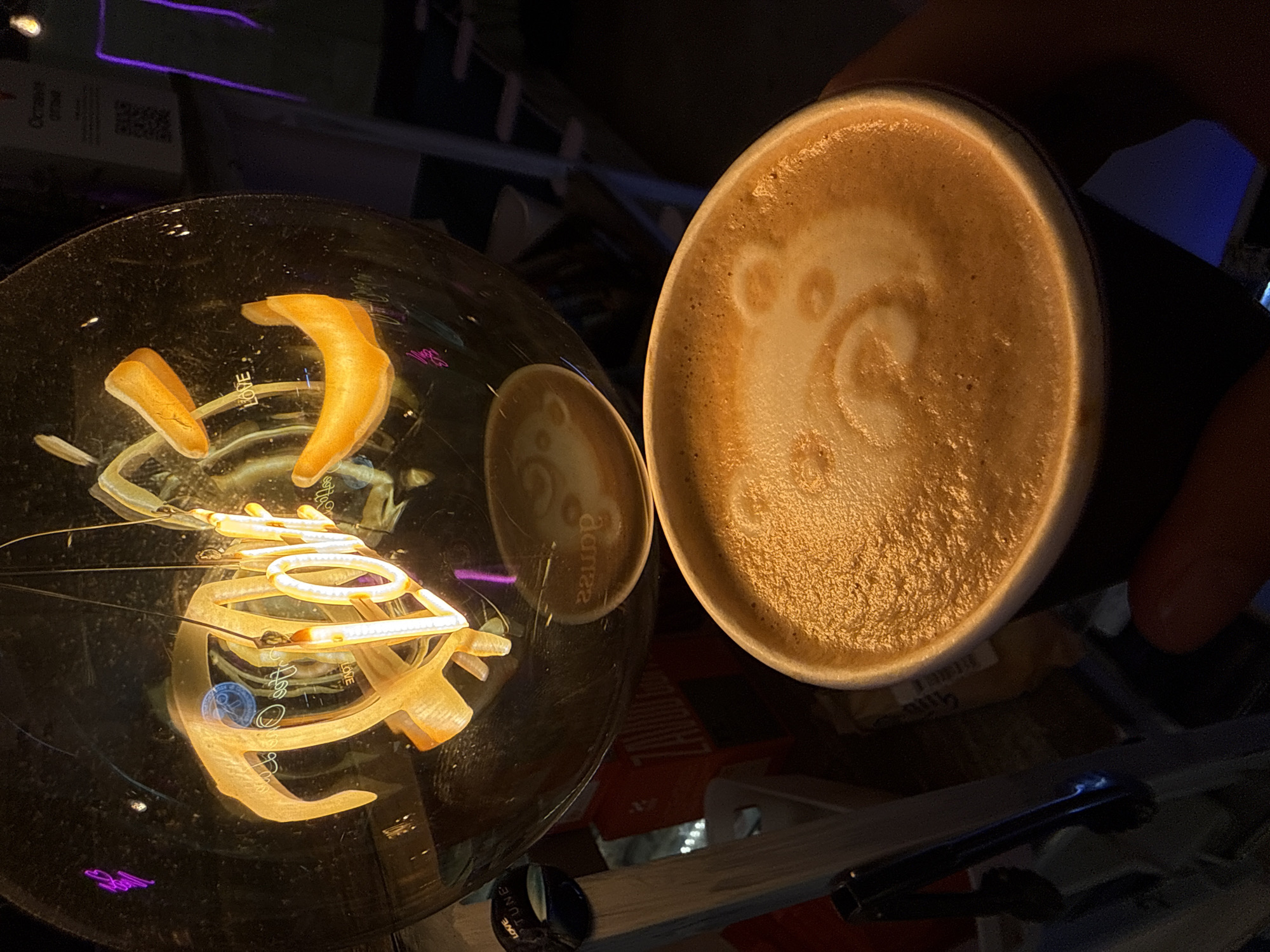 The image shows a close-up of a cup of coffee with latte art and a large, illuminated glass lightbulb in the background. The latte art on the coffee appears to be a bear face. Inside the glass lightbulb, there are illuminated neon-like letters spelling "LOVE" in a stylized font, and also what appears to be a small, red, crescent-shaped object. A purple neon "G" is reflected on the surface of the lightbulb. In the background, there are some shelves with various items, including what looks like boxes and possibly some signage, but the details are indistinct due to the low light and focus. The overall setting appears to be a cafe or bar with moody lighting. The location is not explicitly stated in the image.