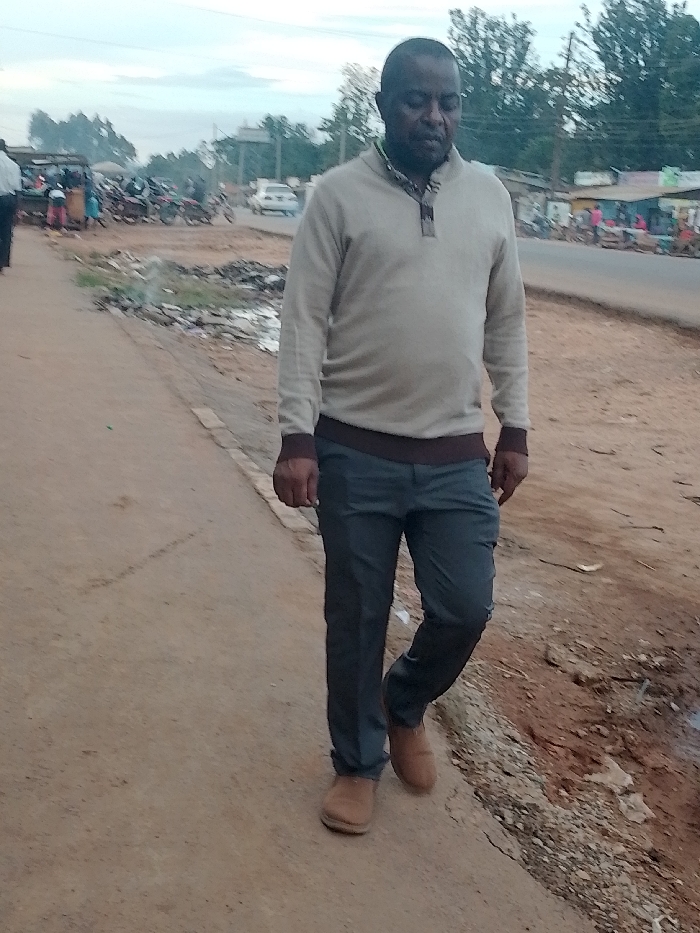 The image shows a man walking on a dirt road in Kivaywa, Kenya. He is wearing a beige sweater, gray pants, and brown shoes. He is walking towards the camera. The road is rough and dusty. There is a paved road in the background, with a few trees and buildings visible. There is debris on the shoulder of the road. There are other people in the background, but they are blurry. The man's face appears to be relaxed but he is looking down. There are no other interactions or activities visible in the image.