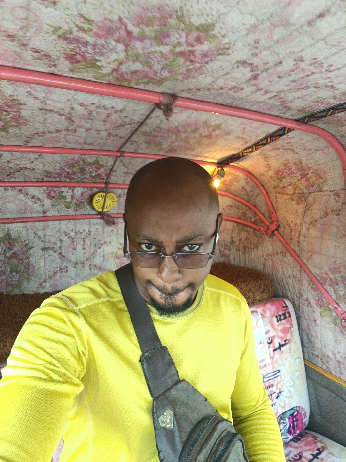 A bald African man with a goatee, wearing glasses and a yellow long-sleeved shirt, poses for a selfie. He is inside a vehicle, possibly a tuk-tuk, with a patterned floral roof and pink metal frame. A brown cross-body bag is slung over his shoulder. A small yellow decorative object hangs from the roof. The vehicle interior appears padded with patterned fabric. A seat is visible to his right.