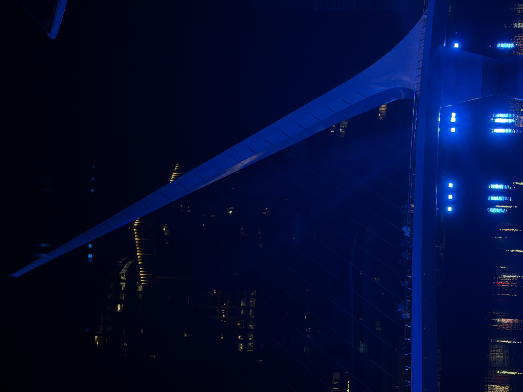 This photograph depicts a bridge at night in Buenos Aires, Argentina. The bridge, illuminated with blue lighting, stretches across a dark body of water. The structure's architecture includes a central pylon with support cables extending diagonally towards the bridge deck. The cityscape is visible in the background, featuring buildings with illuminated windows that reflect in the water. The overall scene is captured from an elevated vantage point, possibly from a nearby building or structure.