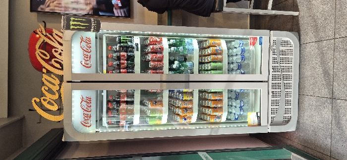 A brightly lit, white, double-door commercial beverage refrigerator stands centrally, fully stocked with various cold drinks. Above the unit, a prominent golden "Coca-Cola" sign with individual letters is mounted, accompanied by a red circular Coca-Cola logo. To the immediate right, a tall, black Monster Energy drink can is depicted on a vertical sign.
The left refrigerator door displays four shelves. From top to bottom, these shelves are stocked with: bottles of Coca-Cola, various Fanta cans (orange, green, yellow, light blue), additional Fanta cans (predominantly orange), and rows of small plastic water bottles with blue labels.
The right refrigerator door also contains four shelves. From top to bottom, these shelves hold: bottles of Coca-Cola and other dark-colored sodas, red Coca-Cola cans, clear or light-colored bottled beverages (possibly Ramune), and assorted Fanta cans (green and orange). Each refrigerator door features a "Coca-Cola" logo near the top. The lower section of the refrigerator is equipped with a grated vent.
The floor is surfaced with light grey square tiles. To the left, a vertical green element, likely a door frame or partition, is visible against a light pink wall. To the right, a person wearing a dark jacket is seated on a dark stool with a silver frame, positioned at a light blue-green counter. Behind the counter, a light beige wall is visible, and a television screen displays a blurred image. The specific location within the city of Villeneuve-d'Ascq, France, cannot be determined from the visual evidence provided.