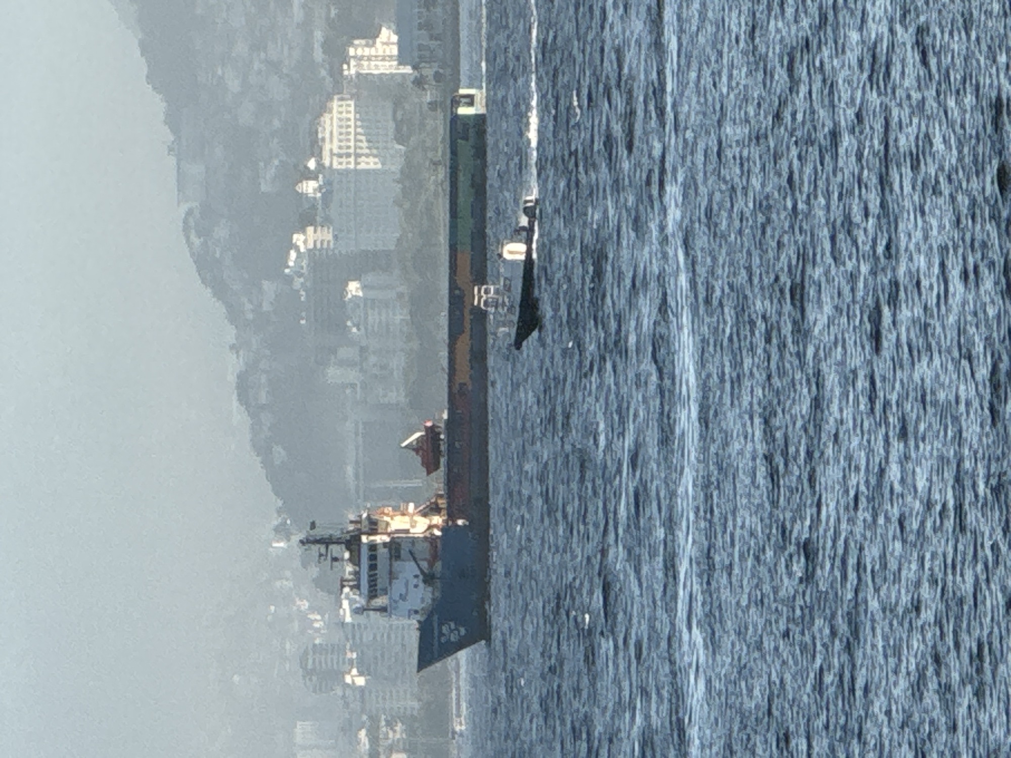 The image depicts a coastal city scene with a large vessel and a smaller boat on the water. In the foreground, the water is choppy with ripples. A dark-colored boat with a white superstructure is moving from left to right, leaving a white wake behind it. In the background, a large ship, likely a cargo or supply vessel, is anchored. This ship is predominantly blue with a white superstructure. Beyond the ships, the cityscape of Niterói, Brazil, is visible. Buildings of various sizes, including apartment complexes and what appear to be older structures, are spread across hillsides. The atmosphere is hazy, obscuring some of the finer details of the city and the horizon. A mountainous terrain forms the backdrop to the city. The lighting suggests it might be daytime, possibly late afternoon, given the soft, diffused light.