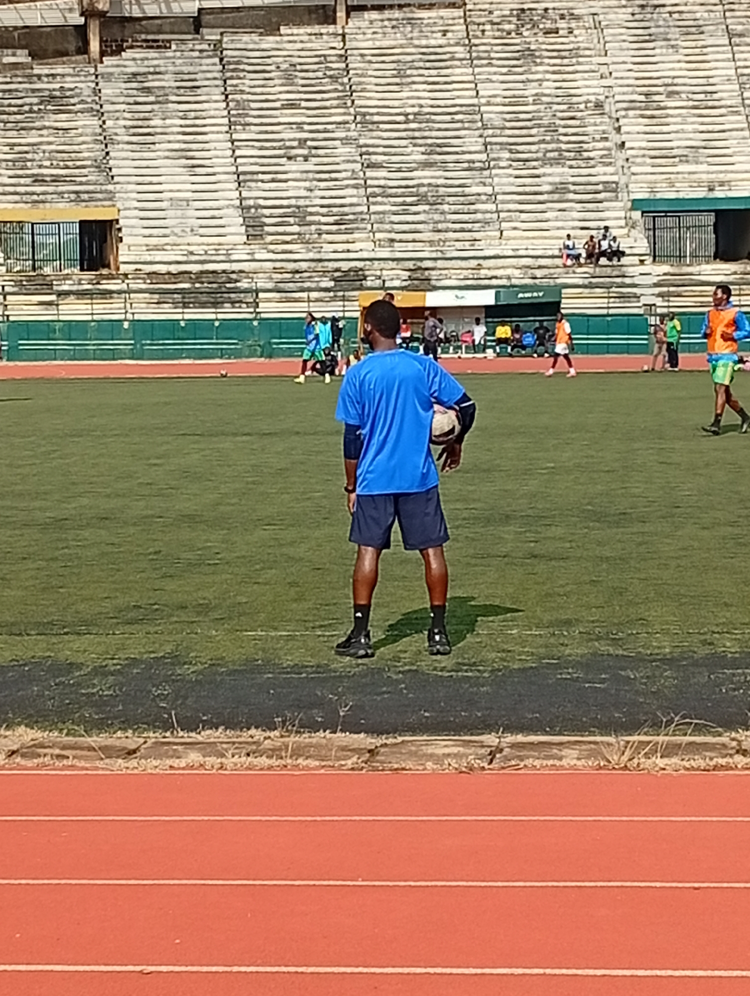 In New Stadium, Nigeria, a male soccer player wearing a blue t-shirt and dark shorts stands on a green turf field, holding a soccer ball. He is facing away from the camera, his back towards the stands. The stands are largely empty, with a few spectators visible in the upper sections. The field has a track running along its edge, colored a distinct reddish-orange. The scene suggests a training or practice session, with other individuals in athletic wear scattered across the field in the background. The stadium appears to be outdoors, with daylight illuminating the scene.
