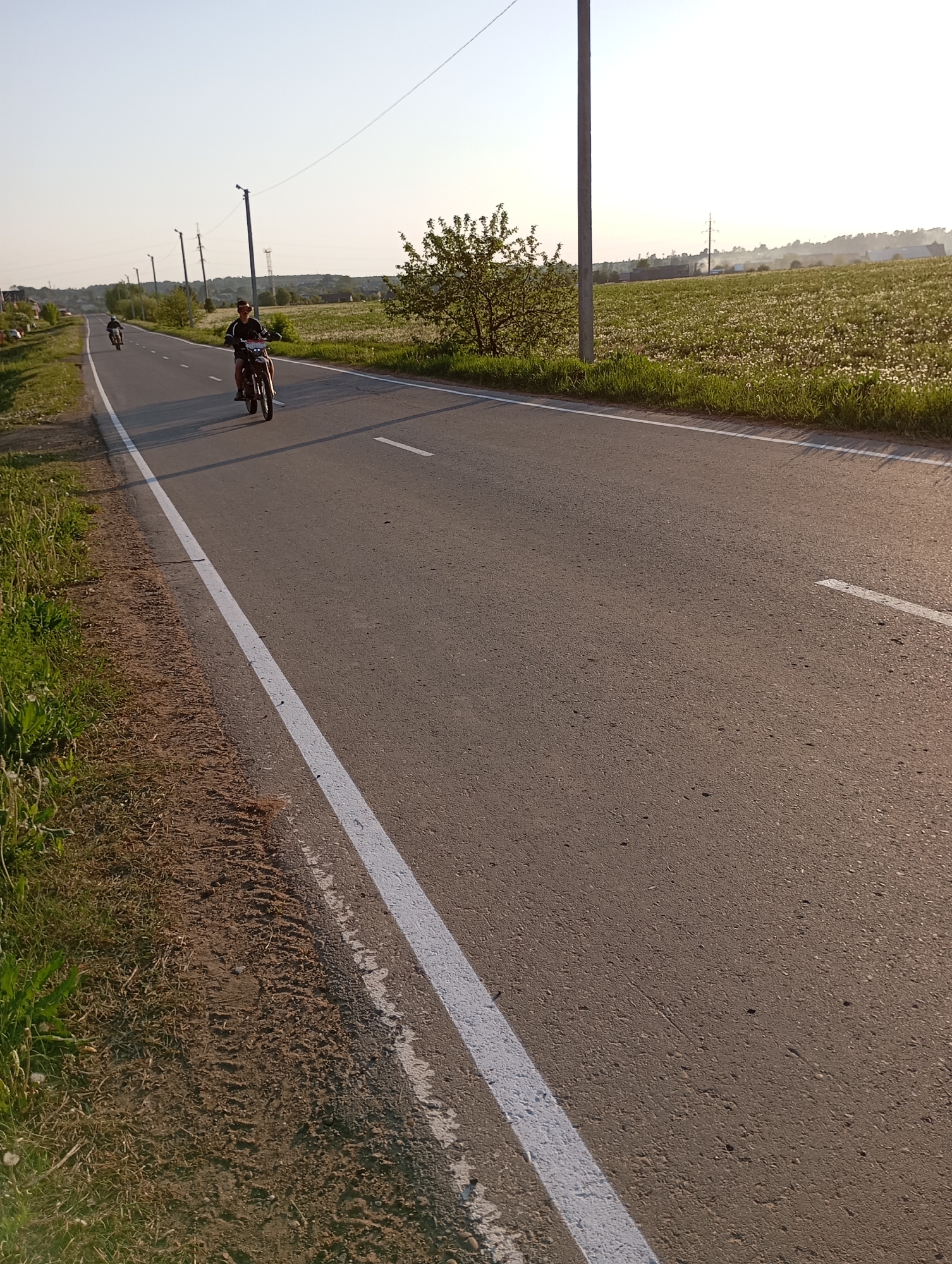 The image depicts a road in Unknown, Unknown, with two people riding motorcycles. The road has a white line marking the edge of the lane. Fields and trees are visible on either side of the road. Utility poles are spaced along the roadside. The sky is clear and bright, suggesting it is daytime.