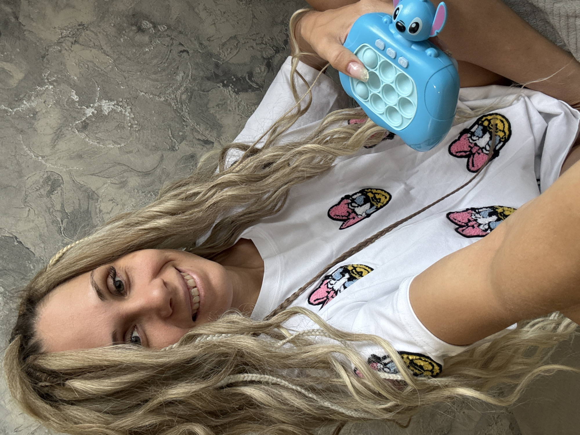 A woman with long, blonde hair with braids is lying down. She is holding a blue Stitch pop-it toy. The woman is wearing a white t-shirt with cartoon designs on it. The background is a grey, textured surface. The location cannot be determined based on the image alone.