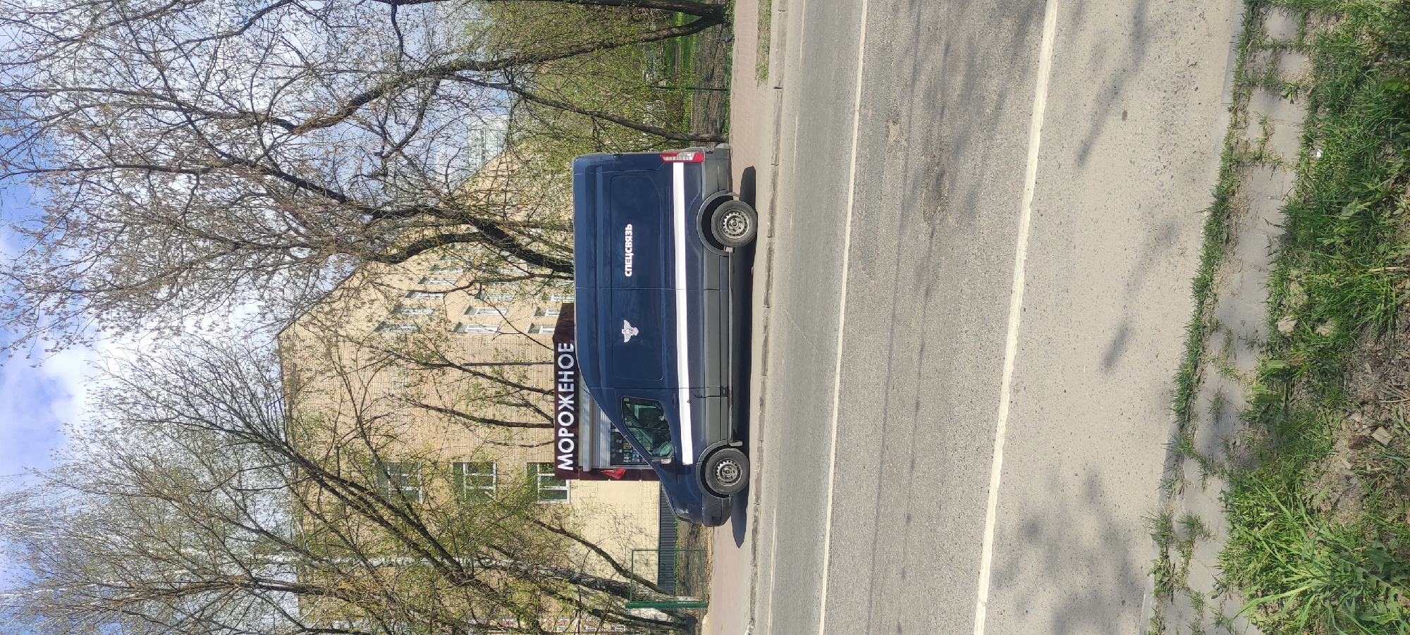 A blue van parked on the side of a road in Fryazino, Russia. The van has white stripes and lettering. The words "Мороженое" (ice cream) are printed vertically on the side of the van. The inscription "Спецсвязь" (Special Communications) is printed next to the emblem of the courier service of the Russian Federation. A building can be seen in the background through tree branches. There is a sidewalk and grass to the right of the road. The road is grey and has white lines painted on it.