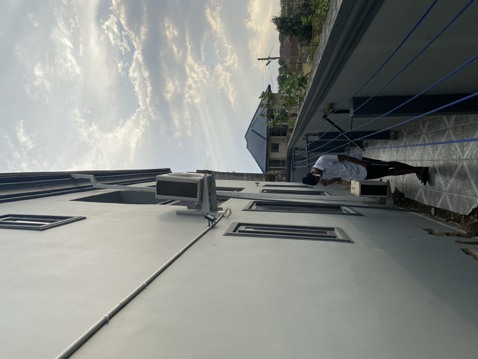 A low-angle perspective shows a light gray building with rectangular windows and an air conditioning unit mounted on the exterior. A man stands on a tiled balcony, looking at his phone. Overhead, a blue sky filled with wispy clouds is visible. The angle is disorienting, as the sky and building are tilted. In the background, a second building with a dark roof and green vegetation is partially visible.