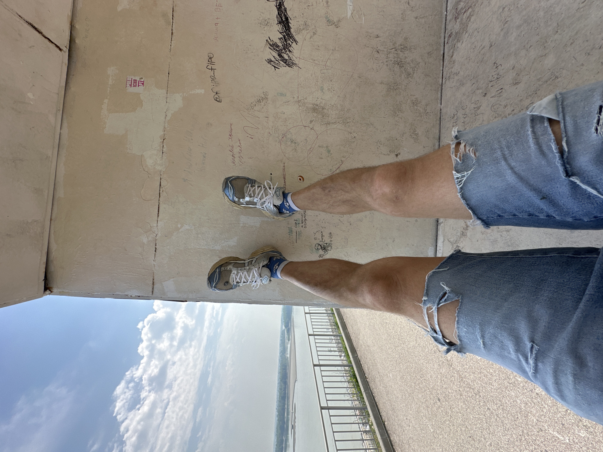 A first-person perspective shot shows a person's legs with ripped denim shorts and athletic shoes as they are suspended against a textured wall. The wall has graffiti and writing on it. To the left of the frame, a railing lines a path, with a body of water and a distant, hazy landscape visible beyond it. The sky above is blue with scattered clouds. The scene appears to be located at the Barnaul embankment in Barnaul, Russia, likely near the observation deck area.