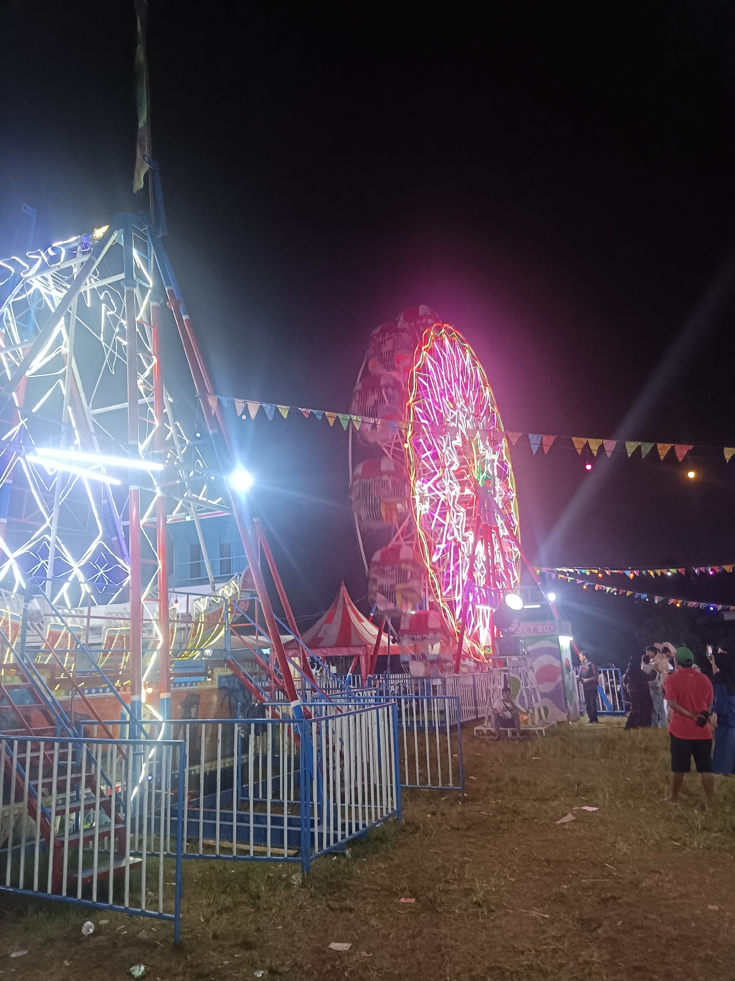 This media file captures a lively night scene at a carnival or amusement park in Depok, Indonesia. The primary subjects are two Ferris wheels, both brightly illuminated with strings of lights. The one on the left is a smaller, more intricate structure with white and blue lights, while the one on the right is larger and glows with vibrant pink and yellow lights.
The scene is set on a grassy field, likely at an outdoor event. A low fence, composed of white and blue bars, encloses a section of the carnival, with stairs leading up to the rides. Small tents, including one with red and white stripes, are also visible. Colorful triangular flags are strung across the scene, adding to the festive atmosphere.
Several people are present, milling around the rides and the ticket booth. Some are standing and watching, while others appear to be waiting in line or exiting the attractions. One person in the foreground wears a red shirt and a green hat, facing away from the camera. The overall mood is one of excitement and enjoyment, typical of a night at a fair.
The time of day is clearly night, with the dark sky serving as a backdrop to the dazzling lights of the rides. There are no obvious weather cues, but the ground appears to be dry grass. A sign reading "TICKET BOX" is visible on a booth near the larger Ferris wheel. The image conveys a sense of fun and community engagement, with the bright lights and the presence of many people indicating a popular event.