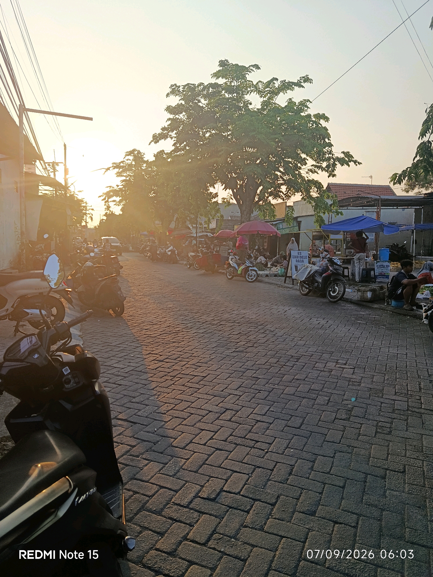 The image depicts a street scene in Keboansikep, Indonesia, at sunrise. The street is paved with interlocking brick pavers, and parked motorcycles line both sides. In the foreground, several motorcycles are visible, some with reflective surfaces. On the right side of the street, market stalls are set up under awnings, with vendors and customers interacting. One stall has a sign with Indonesian text and an image of an ice product. Further down the street, more motorcycles are parked, and a white van is visible in the distance. The sun is low in the sky, casting long shadows and illuminating the scene with a warm, golden light. Trees with lush green foliage are present throughout the scene. The overall atmosphere is that of a typical morning market activity.