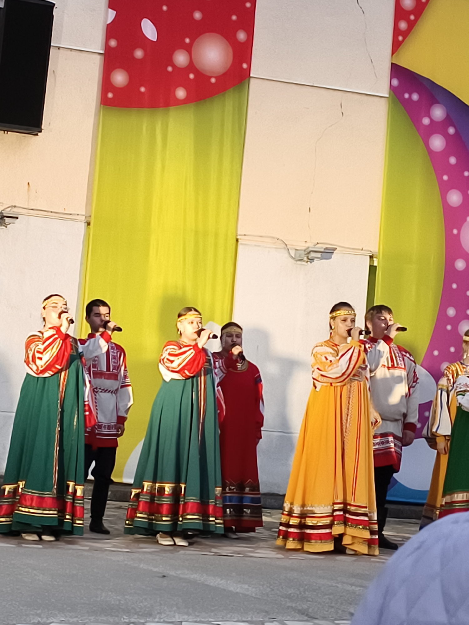 This image captures a group of performers on a stage, likely during a cultural or folk music event. Six individuals are clearly visible, with more partially shown. Three women are prominent, two wearing vibrant deep green, long-sleeved dresses (sarafans) with red, white, and gold trim, and one in a bright yellow/orange dress with similar elaborate patterns and a red and gold striped design. All three wear decorative headbands and are holding microphones, appearing to sing with focused expressions.
Accompanying them are three men. Two are dressed in white long-sleeved shirts (kosovorotkas) with intricate red and black embroidery, paired with dark pants. One man in a red top is also visible further back. They are also holding microphones and seem to be part of the vocal ensemble. The performers' traditional attire, particularly the sarafans and kosovorotkas, strongly suggests a Slavic or Russian cultural performance.
The setting appears to be an outdoor stage or an open-air venue. The backdrop behind the performers is colorful and abstract, featuring large sections of red with white polka-dots (resembling a mushroom cap), interspersed with bright lime green fabric panels and a curved pink/purple section also adorned with white circles. Plain white wall segments are visible between and above these decorations, showing minor cracks, some discoloration, and exposed electrical conduits or wires. A black speaker or monitor is mounted on the wall in the top-left corner. The ground is a uniform gray, likely asphalt or concrete. The lighting suggests it is daytime or early evening. No text is visible in the image.