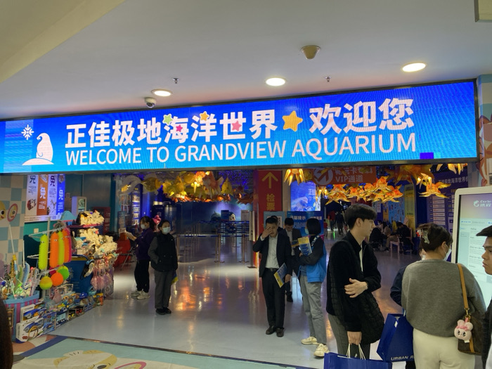 This indoor photograph captures the brightly lit entrance of the Grandview Aquarium, located in Guangzhou, China. A large, blue LED display sign dominates the top of the frame, featuring the Chinese text "正佳极地海洋世界★欢迎您" (Grandview Polar Ocean World ★ Welcome), with "WELCOME TO GRANDVIEW AQUARIUM" prominently displayed in English beneath it. The sign also includes a graphic of a penguin and star motifs.
Beneath the main sign, golden fish-shaped decorations are suspended, leading into the aquarium's main entry point. On the left side of the image, a retail display area showcases a variety of colorful toys and merchandise, including plush items and novelty goods. Several people are visible throughout the scene, indicating an active public space. Two individuals, one wearing a face mask, stand near the merchandise section. Towards the center, a man in a dark jacket carries a shopping bag with "URBAN" printed on it, while other visitors are gathered on the right, with one person seemingly interacting with a digital information kiosk.
Smaller signs are also present: "免" (free/exempt) is visible near the merchandise, and signs indicating "VIP通道" (VIP Channel) and "检票" (Ticket Check) are seen closer to the entrance. The overall atmosphere suggests a normal operating day at a popular tourist attraction, with people either entering, browsing, or waiting to access the aquarium. The presence of face masks on some individuals suggests either health precautions or the timeframe of the photograph.