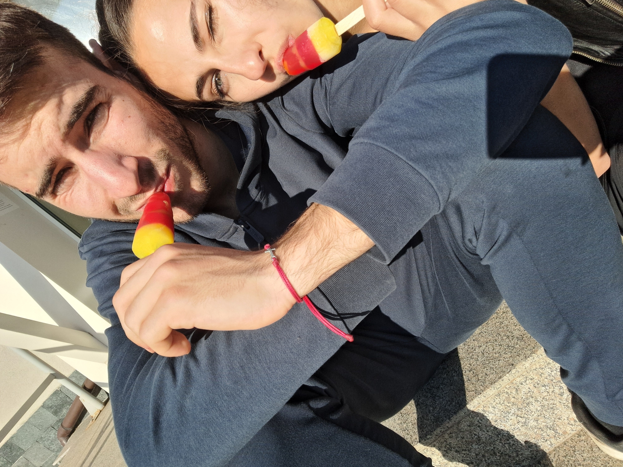 A medium-shot captures two individuals, a man and a woman, seated outdoors and embracing each other while sharing ice pops. The man, with dark hair and a beard, holds a red and yellow ice pop to his mouth. He wears a dark blue hooded sweatshirt and matching pants, and a red bracelet with a silver cross on his left wrist. The woman, with dark hair pulled back, rests her head on the man's shoulder and also holds a red and yellow ice pop to her mouth. She is wearing a dark-colored top. The background shows a light-colored stone surface and a white railing. The scene is bathed in sunlight, creating shadows across the ground.
