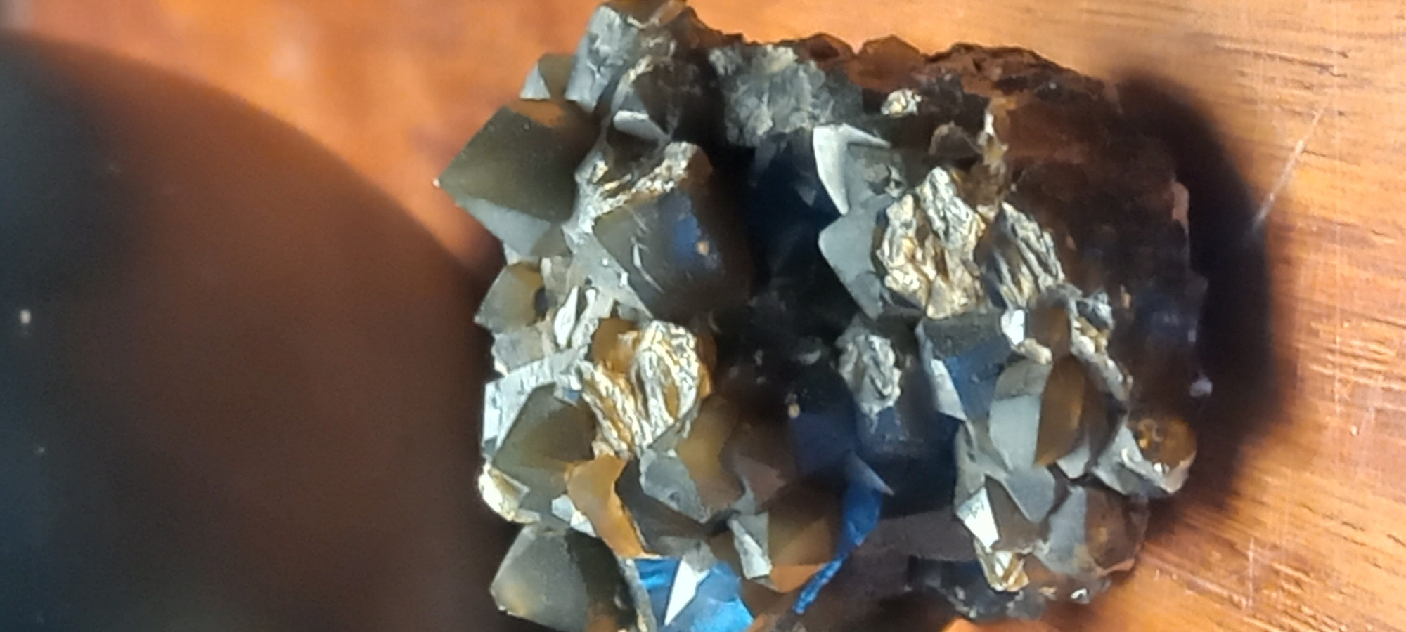 A close-up shot captures a mineral specimen composed of numerous dark, crystalline structures intergrown with a lighter, metallic-looking mineral. The primary crystals are dark, appearing black, deep grey, or dark brown, with discernible facets and sharp edges characteristic of well-formed crystals. Some areas of these dark crystals exhibit a subtle blueish reflection or tint, possibly due to lighting or inherent mineral properties.
Interspersed among these dark crystals, and in some areas appearing to coat them, are clusters of a brighter, more irregularly textured mineral. This mineral possesses a distinct metallic luster, appearing golden-bronze or silvery, and contrasts sharply with the darker crystals. It has a more granular or flaky appearance compared to the geometric forms of the main crystals.
The specimen rests on a warm-toned, reddish-brown wooden surface, which forms the background on the right and upper portions of the image. On the left side, a large, blurred, dark, curved object is partially visible, suggesting an indoor setting with other objects in the immediate vicinity.
The lighting appears directional, creating highlights on the crystal faces and metallic portions, and casting subtle shadows that emphasize the three-dimensional structure of the mineral cluster. There is no human activity depicted, nor is any specific time of day or weather discernible. No text is visible in the image. The location is contextualized as Kryvyi Rih, Ukraine.