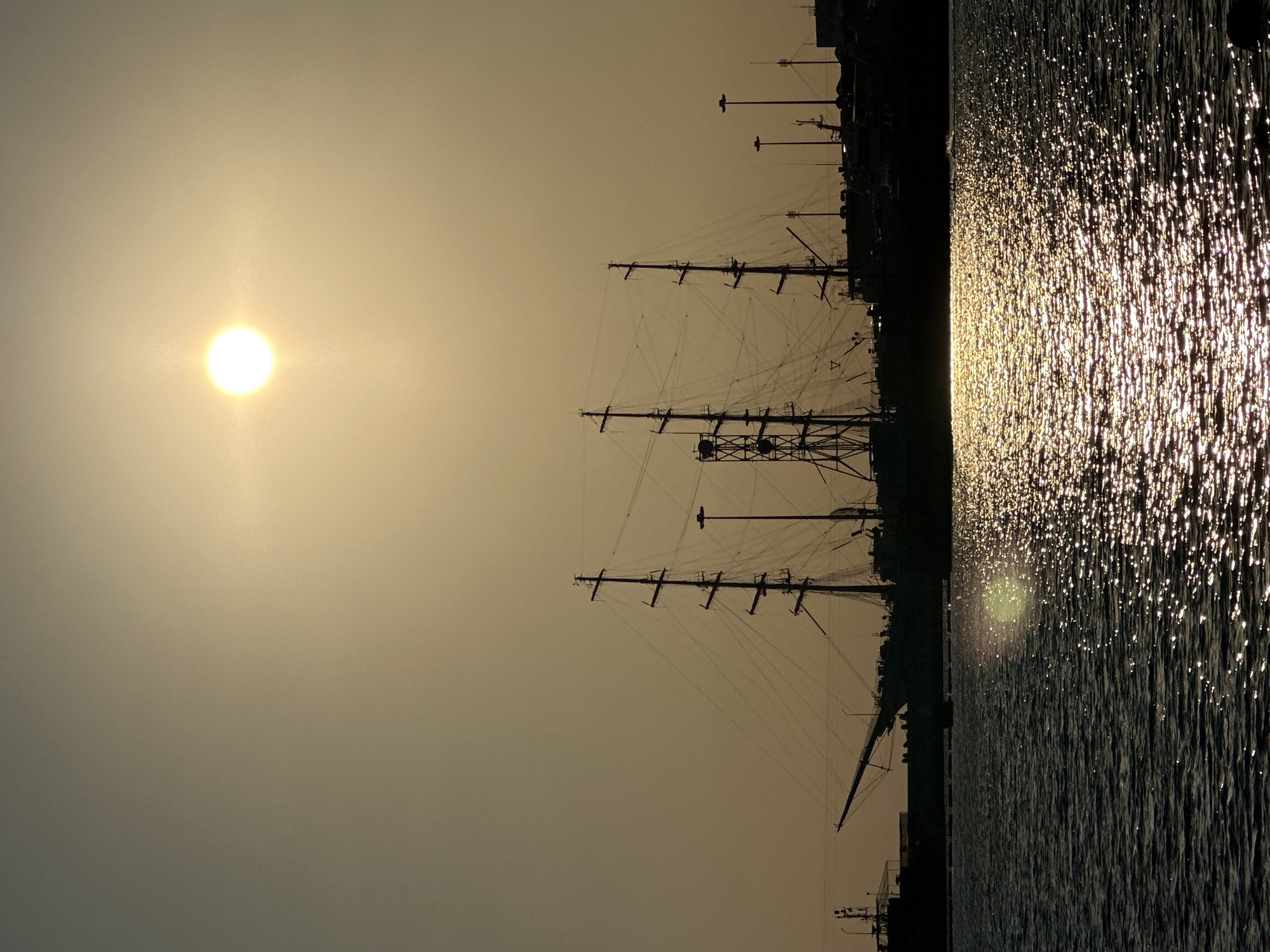 The image captures a serene sunset scene at the Sochi port in Russia. The sun, a brilliant orb, hangs low in the hazy sky, casting a warm, golden glow that reflects off the rippling water. Silhouetted against the vibrant sky is a majestic tall ship, its masts and rigging intricately detailed. Several other vessels and harbor structures are visible in the background, also rendered as dark shapes against the fading light. The water surface is alive with shimmering highlights, a testament to the sun's descent. The overall atmosphere is one of tranquility and maritime beauty.