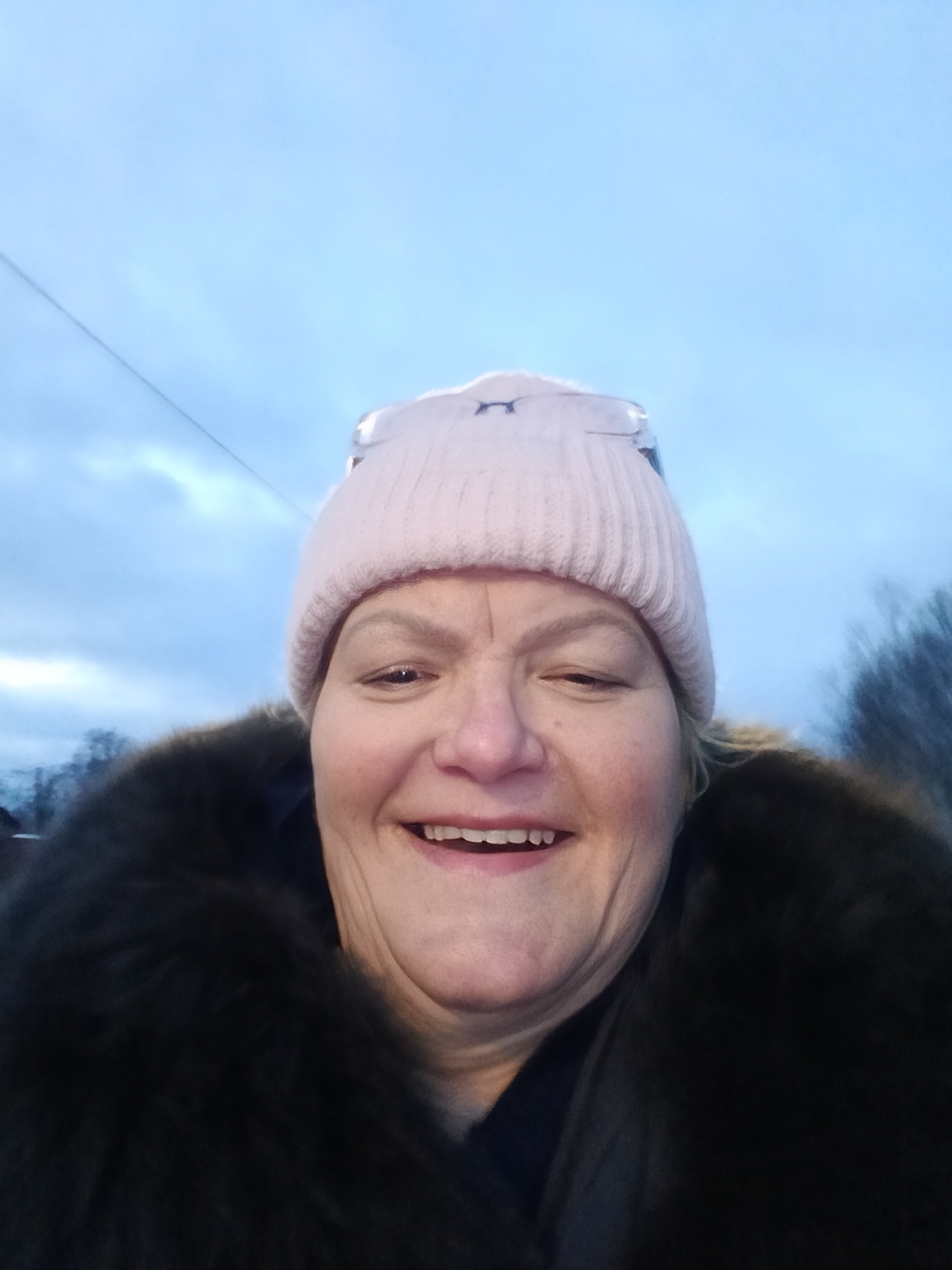 A woman is taking a selfie outdoors. She is wearing a pink beanie and a dark, fur-trimmed coat. She is smiling. The background includes a cloudy sky and some blurred trees. The location is likely Krasny Mayak, Russia.